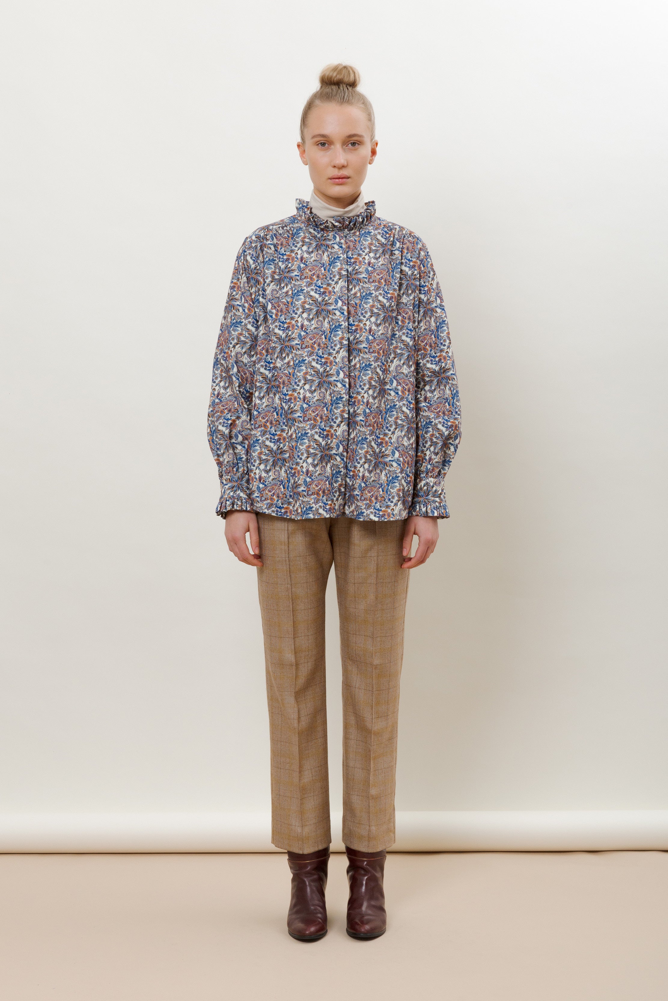 Woman in multi-colored flower patterned shirt with frilled collar and long sleeves and brown pants