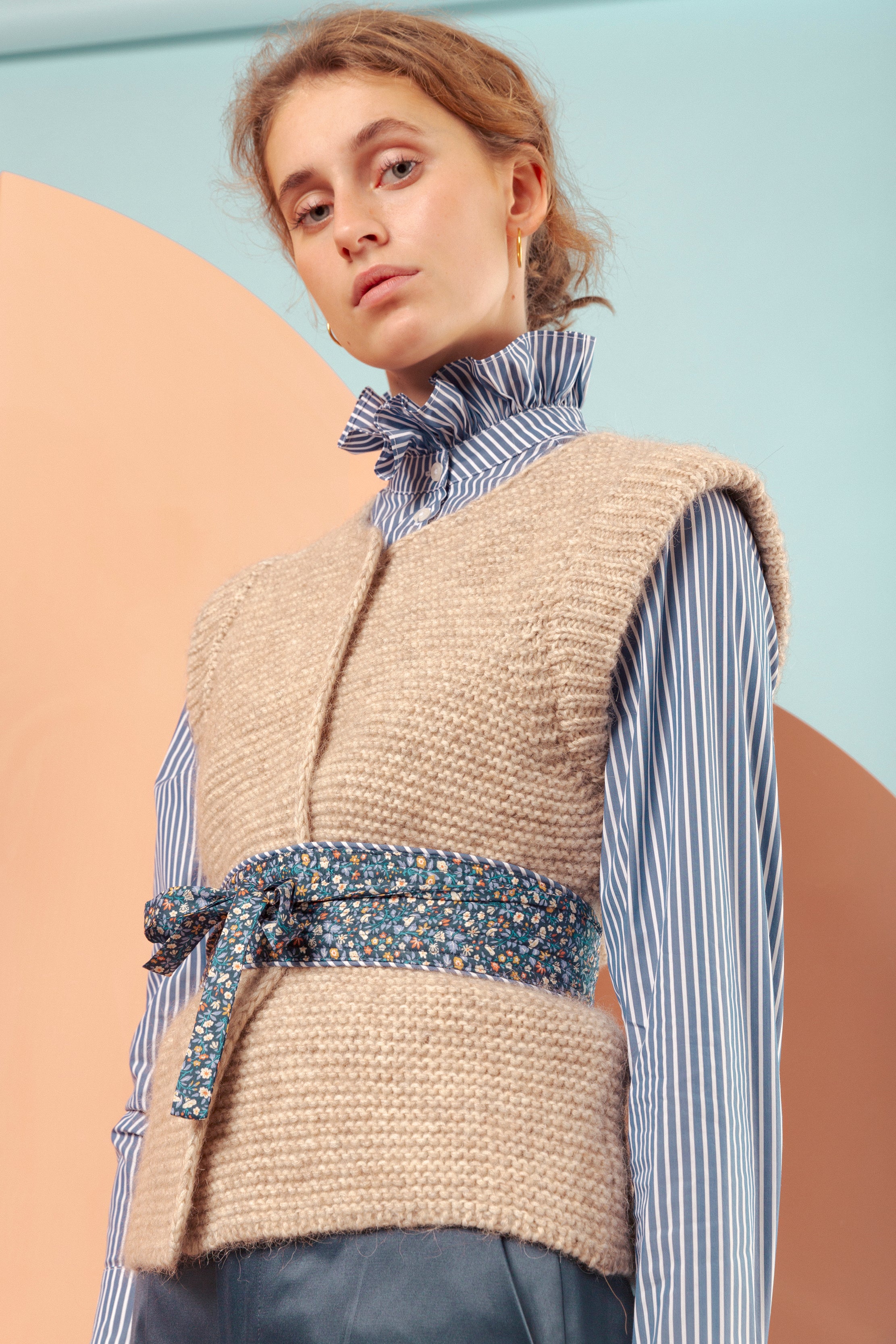 Woman in blue and white striped shirt with frills, beige knitted wool vest, flower patterned belt and blue pants