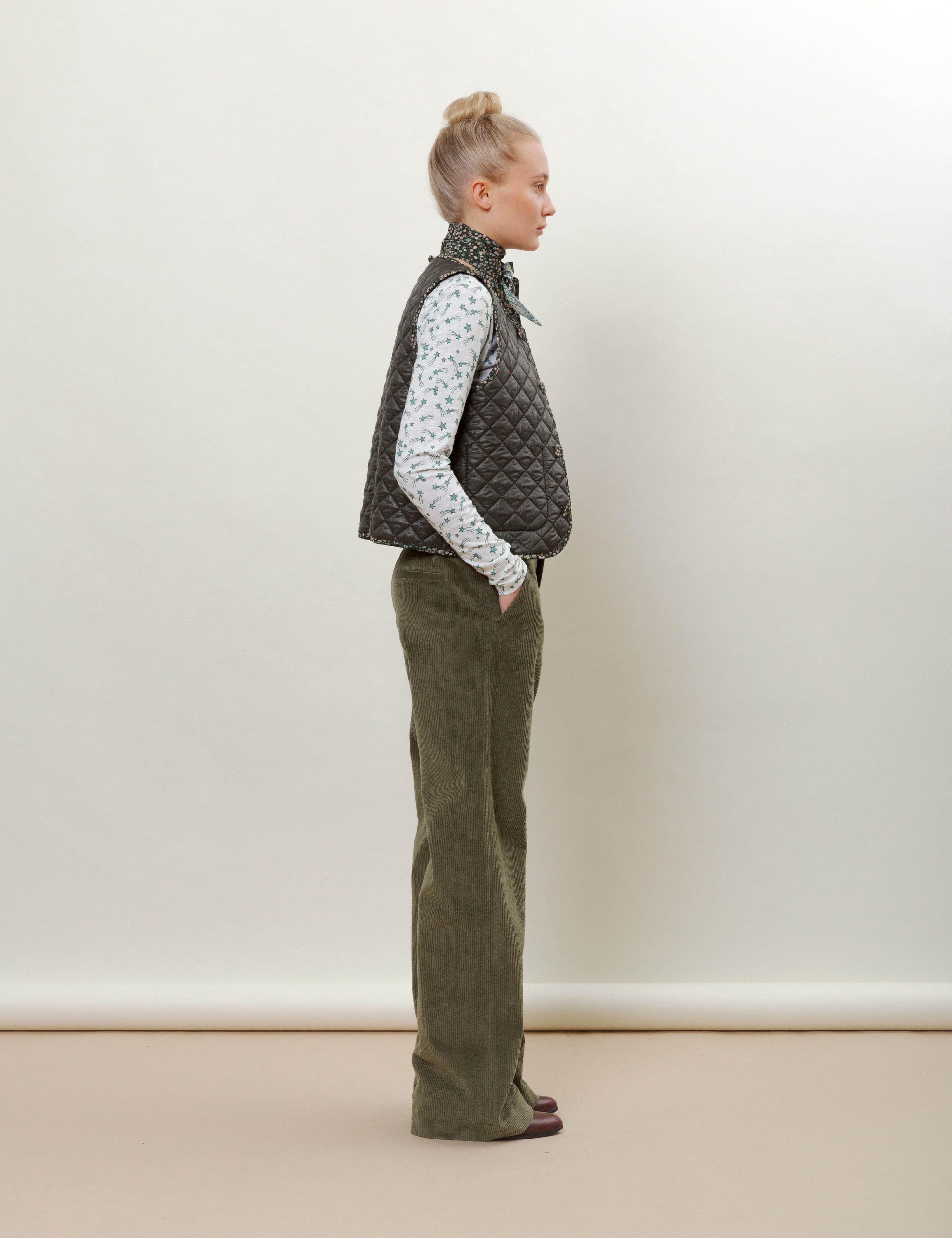 Woman in green corduroy pants, green vest, patterned t-shirt and scarf