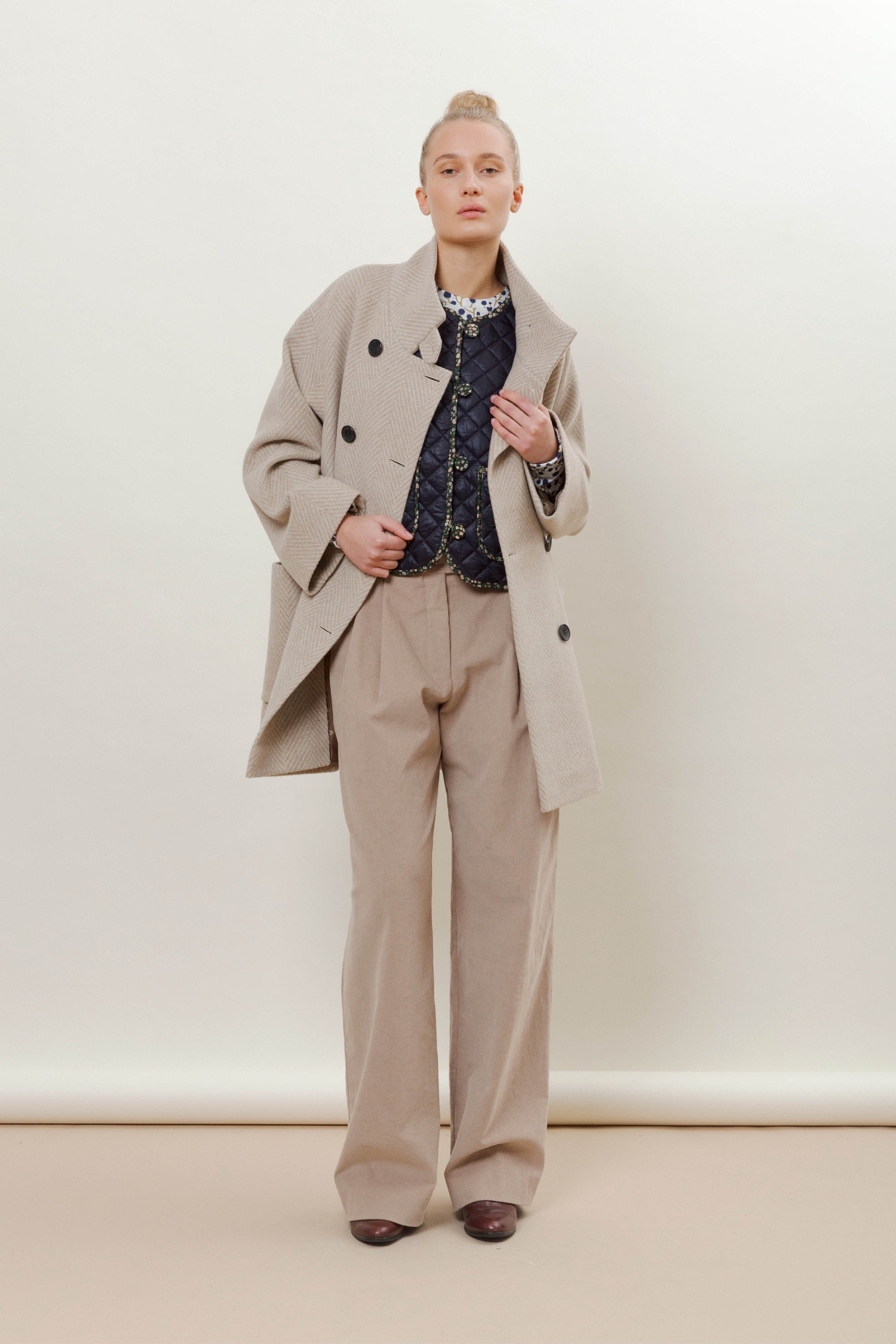 Woman wearing beige herringbone patterned wool jacket with black buttons, pockets and stand collar, beige pantd and blue vest