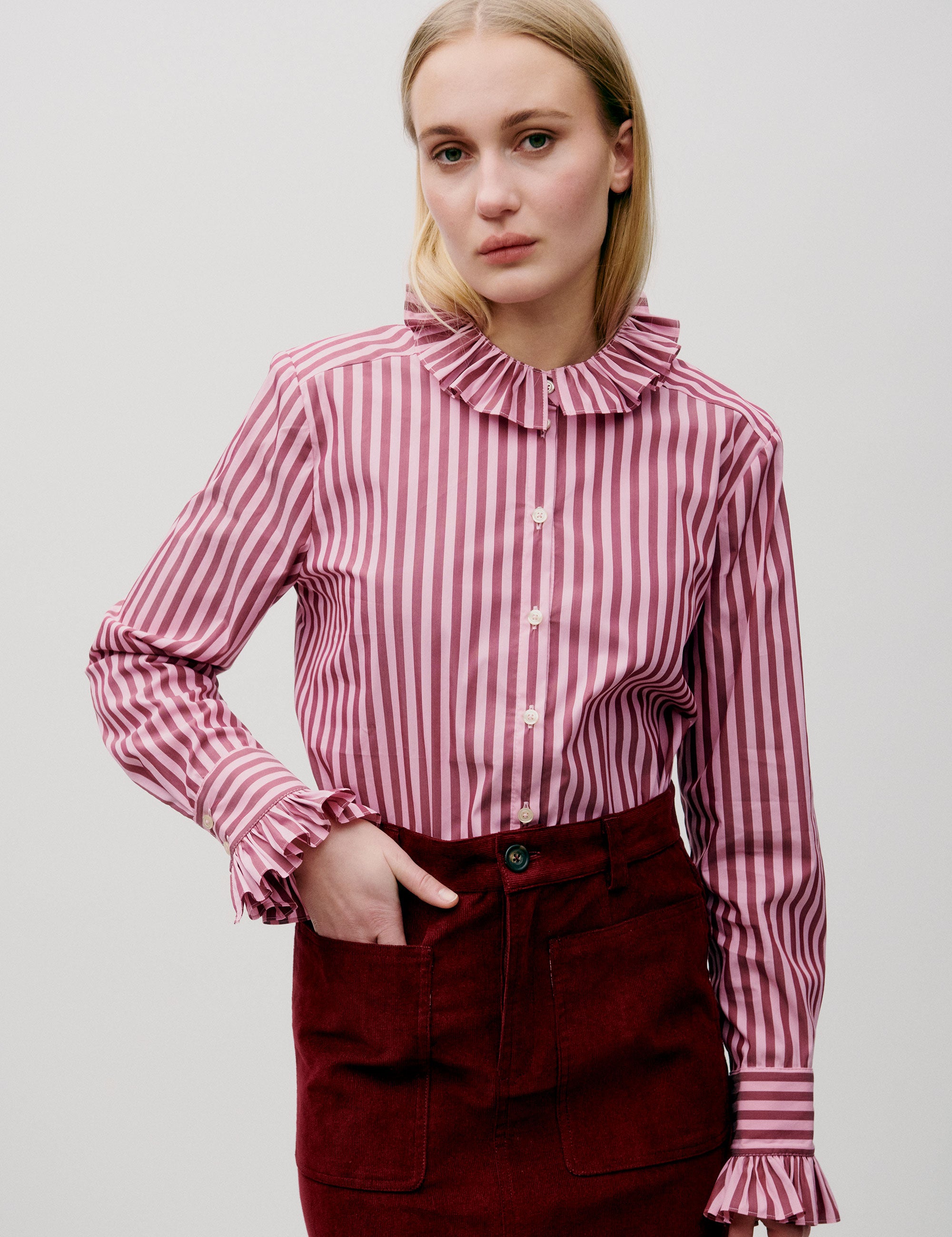 Woman wearing a pink and white striped blouse with a high collar and red skirt on a light gray background