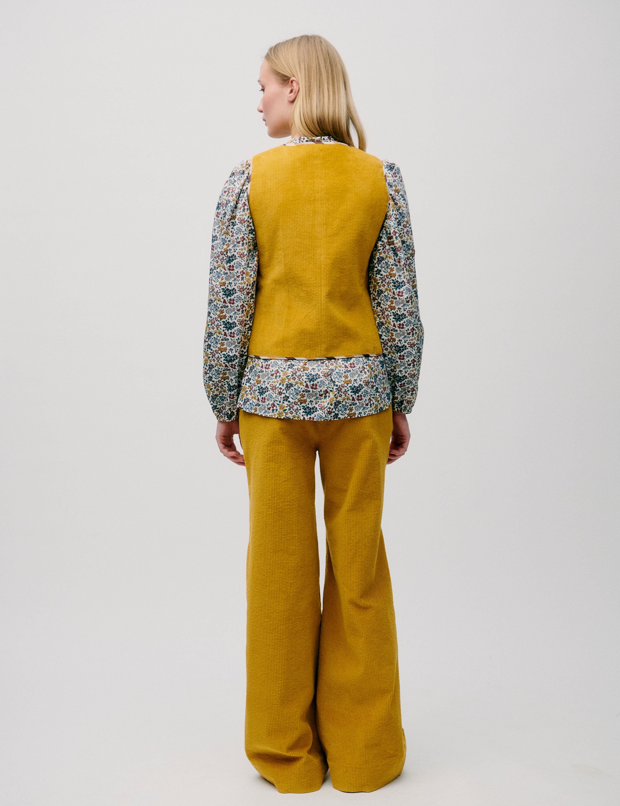 Woman seen from behind in a yellow vest and trousers, with a floral blouse underneath.
