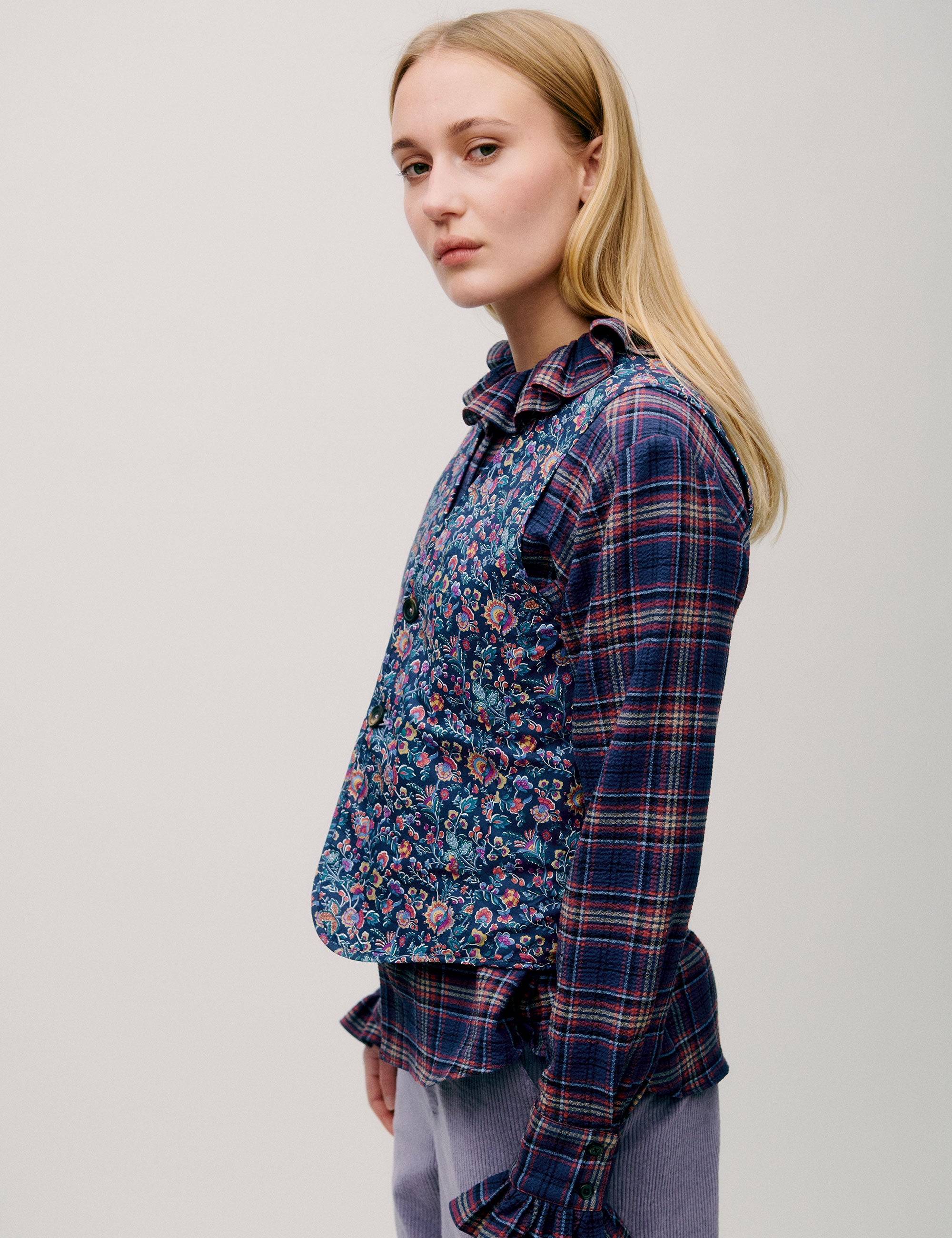 Side view of dark check blouse with ruffle details, floral vest and lilac trousers