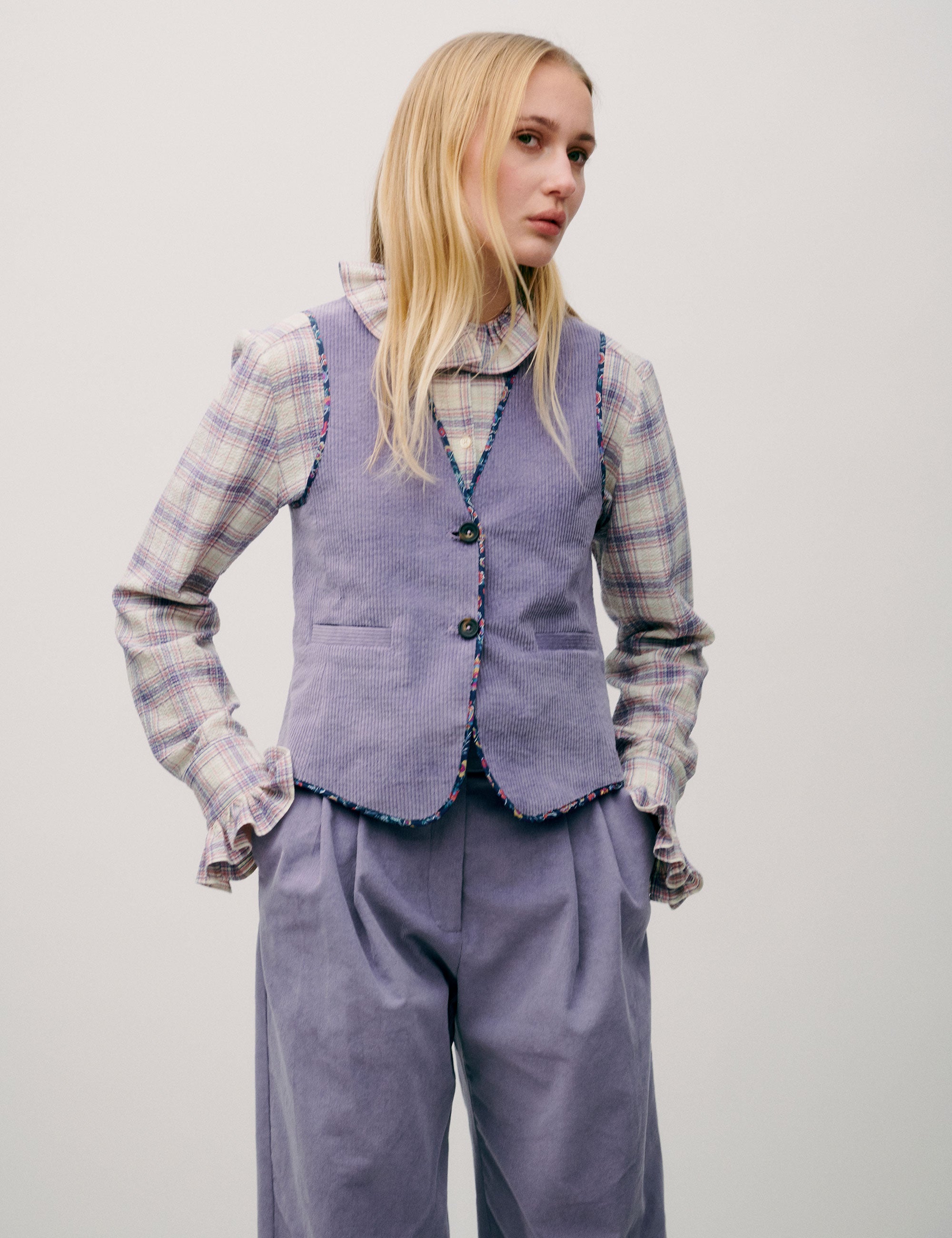 Woman wearing a checked blouse with ruffle collar under a striped lilac vest and high-waisted wide lilac corduroy trousers