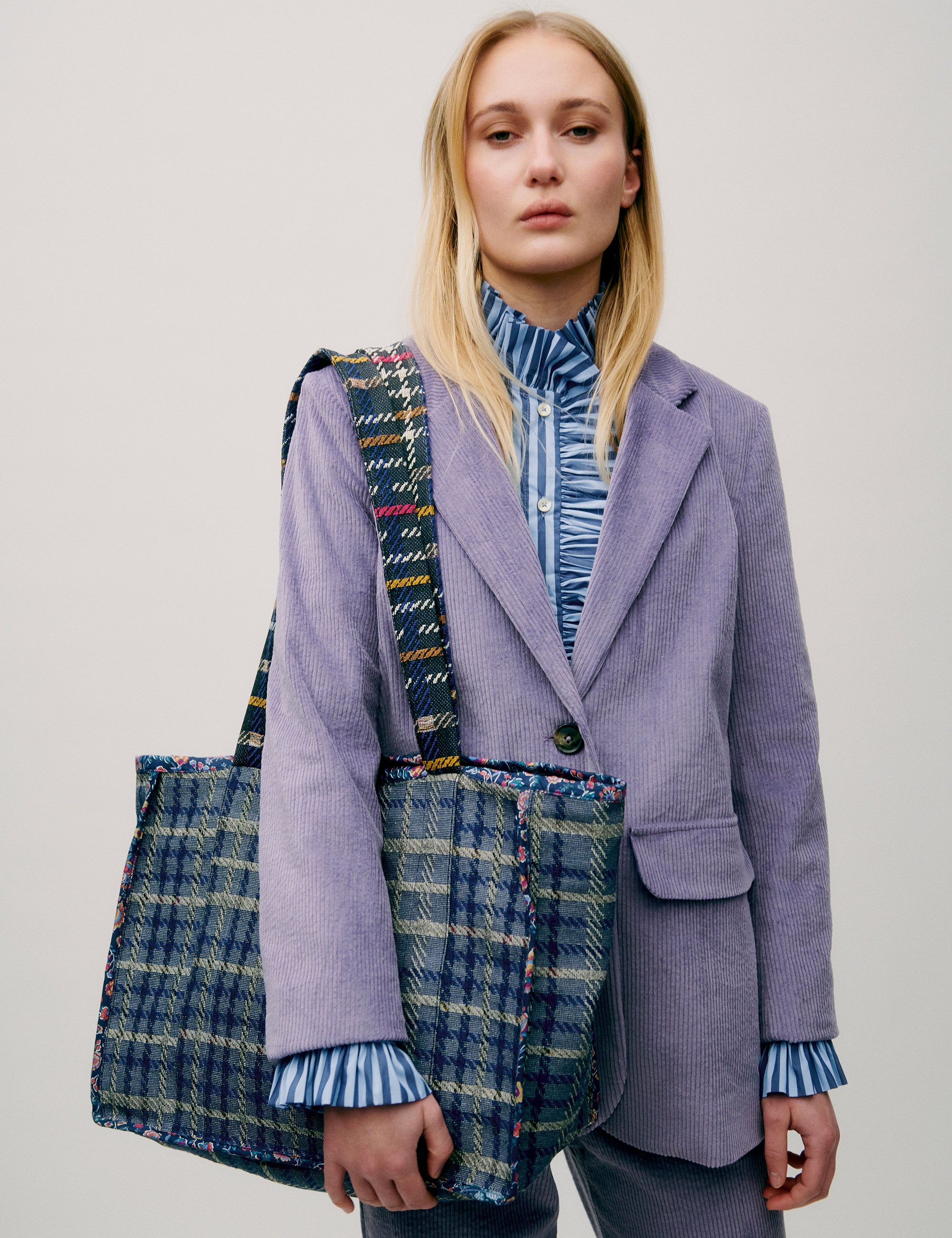 Woman in lavender colored corduroy pants and jacket and blue striped shirt with frills carrying a large check patterned cotton tote bag