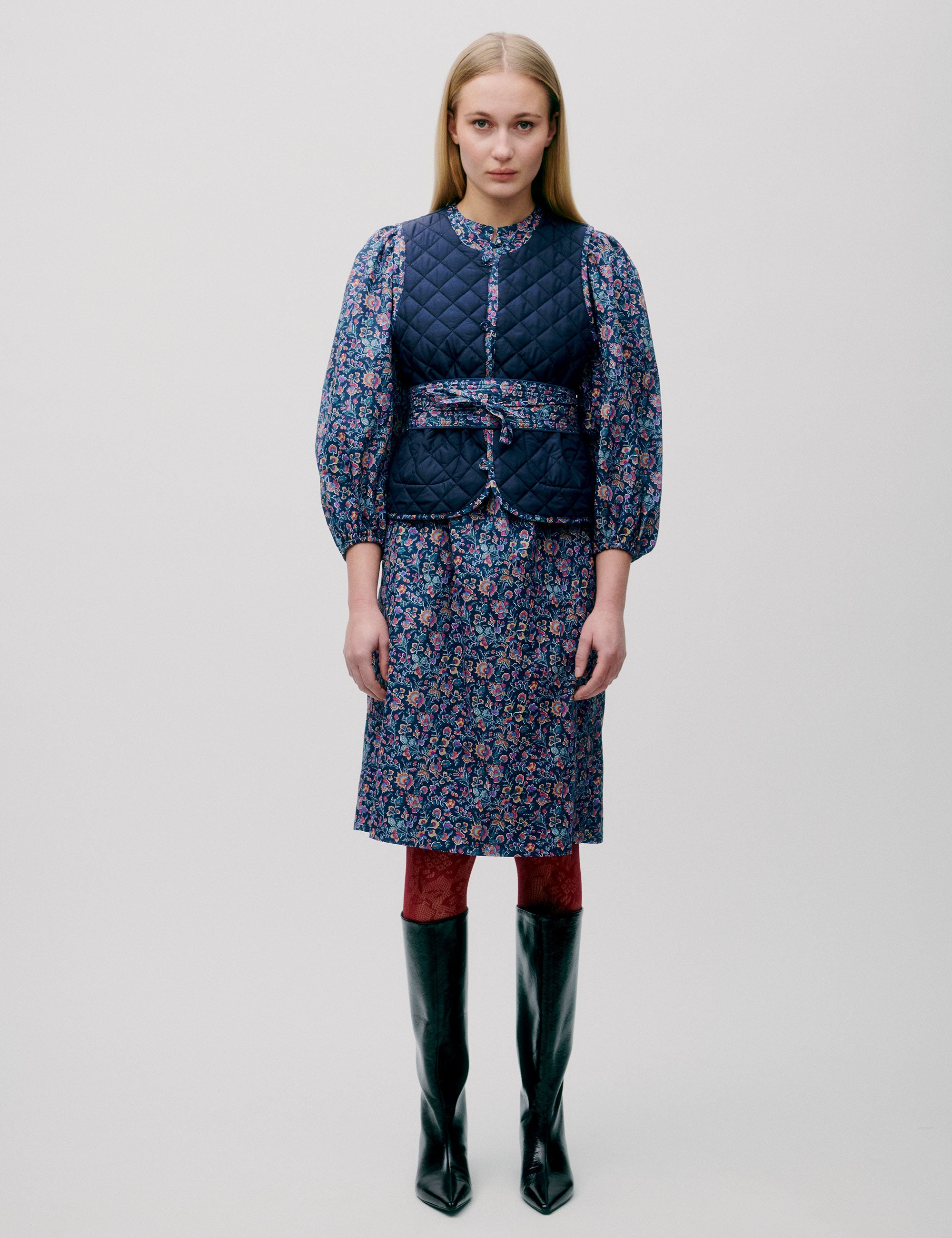 Woman in pink and blue flower patterned short dress with long sleeves, quilted vest and matching cotton belt
