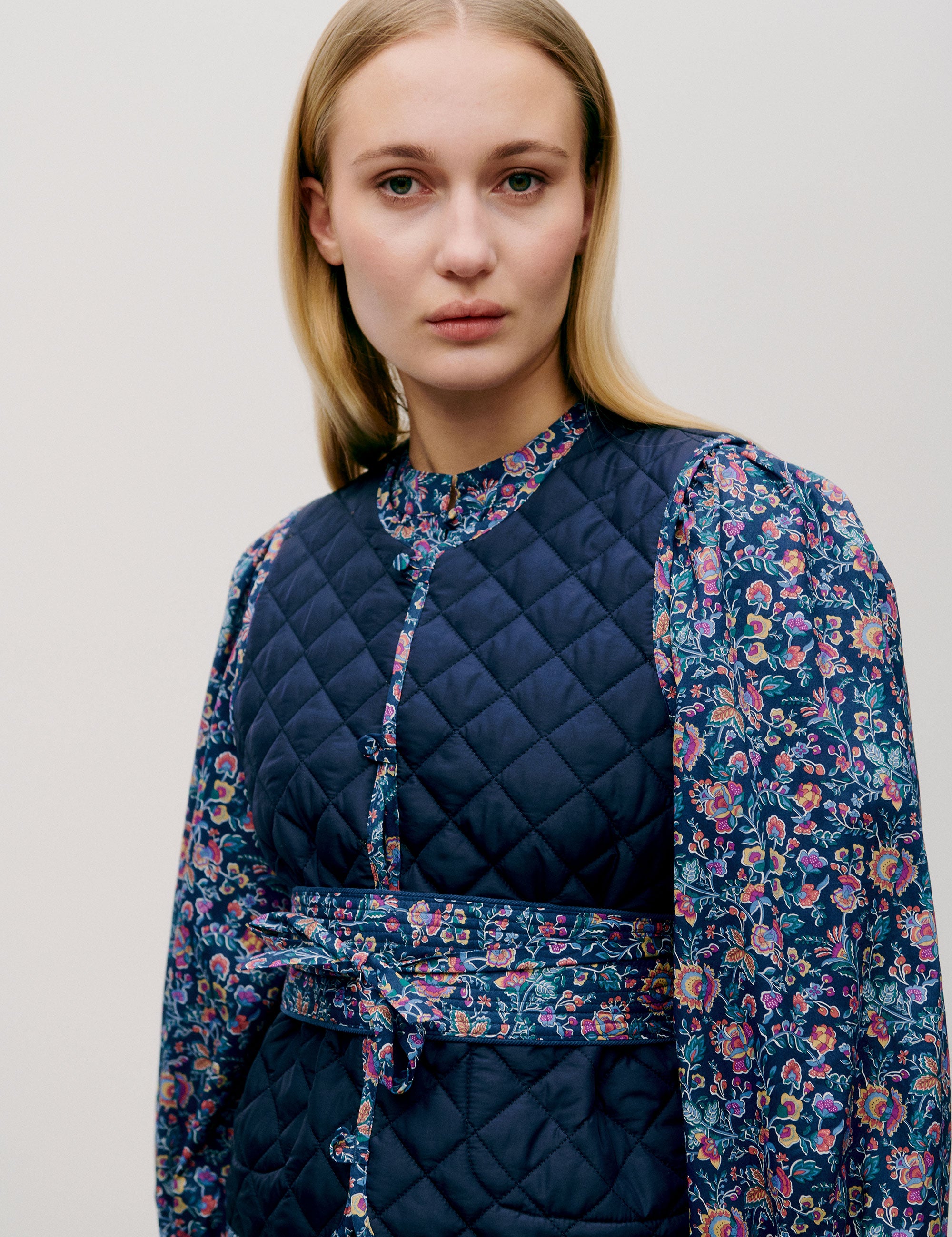 Woman in pink and blue flower patterned short dress with long sleeves, quilted vest and matching cotton belt