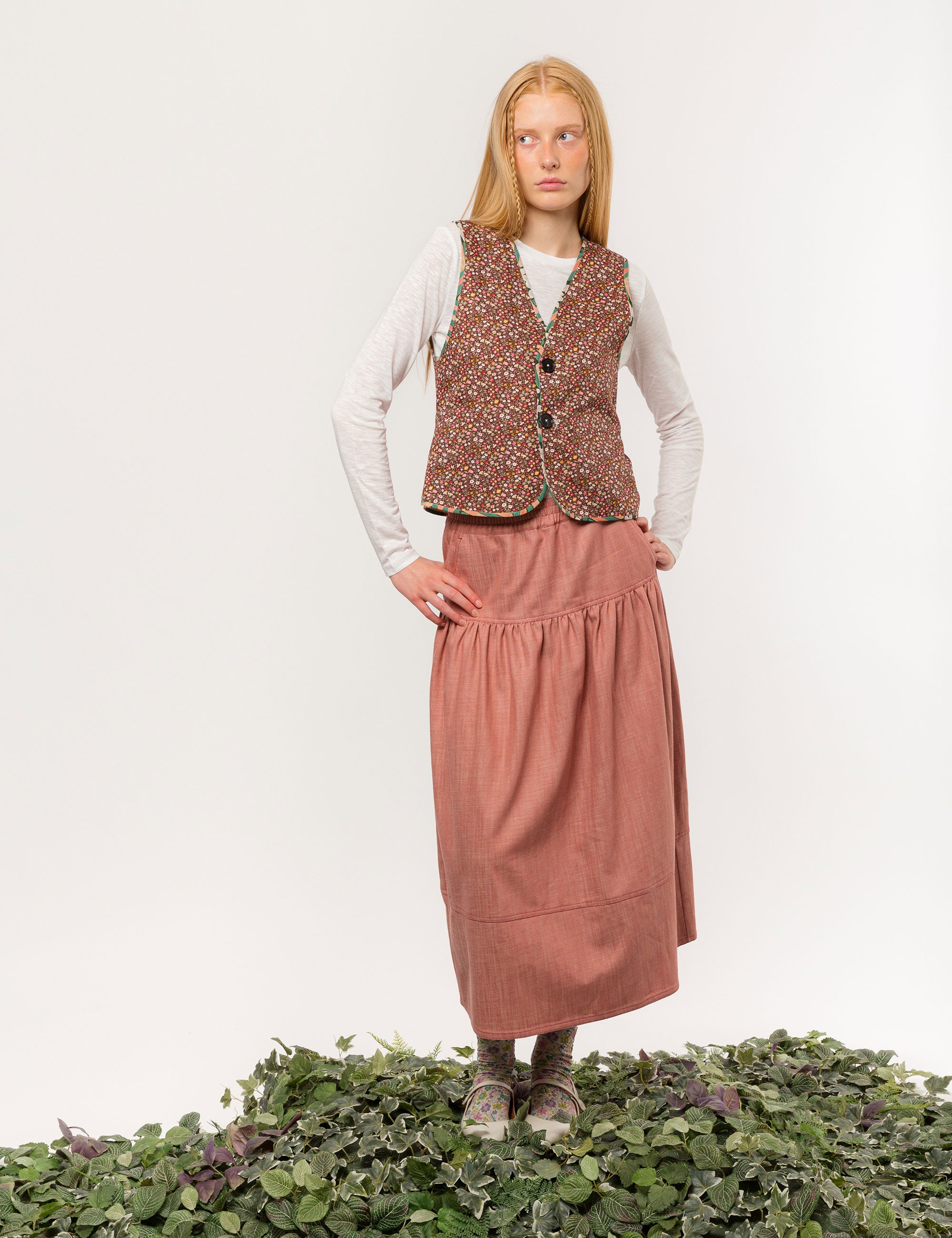 Woman in reversible cream colored vest with buttons, pockets and floral print pipings, white long-sleeved t-shirt and pink maxi skirt