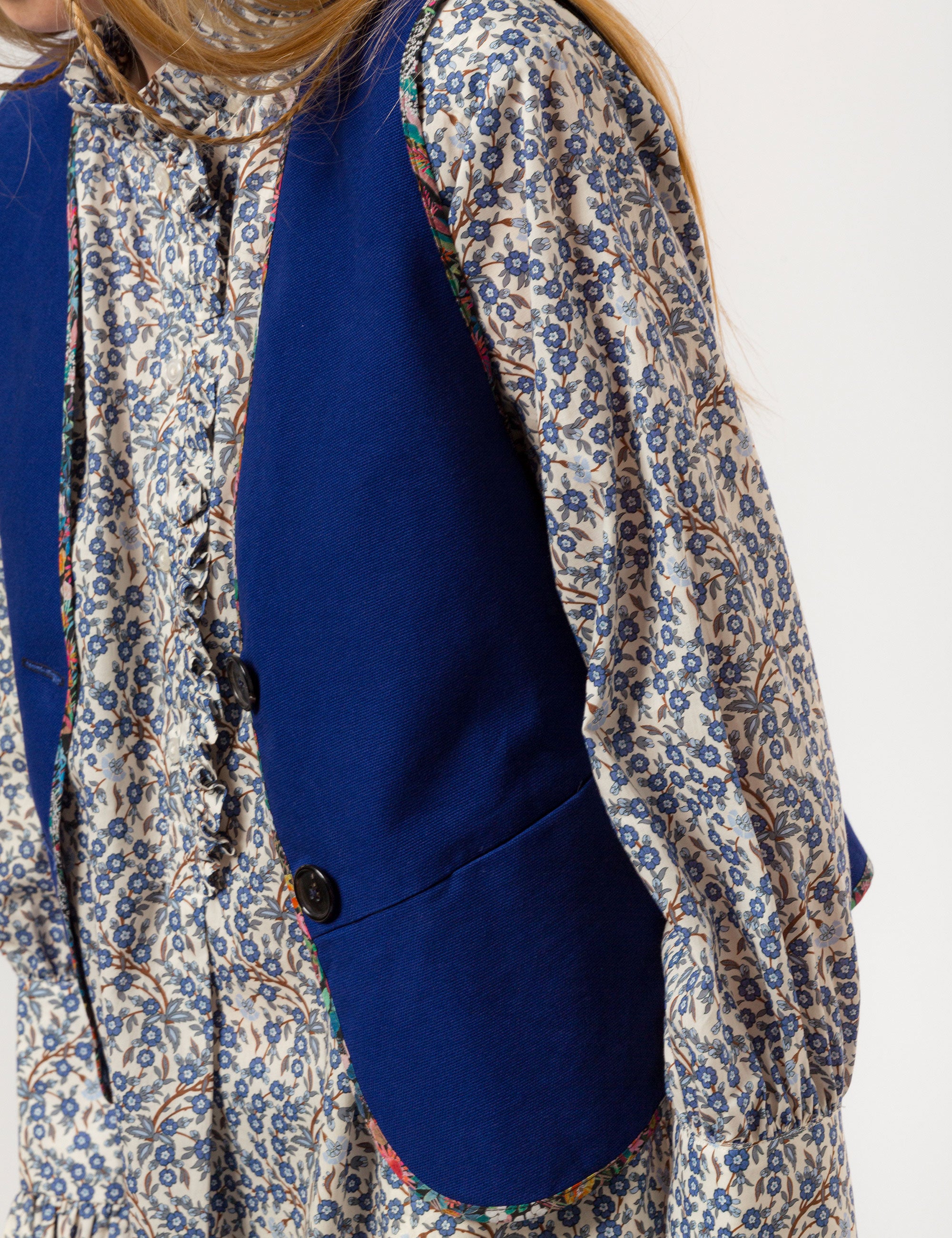 Woman in long sleeved white and blue floral print shirt and blue vest with buttons and pockets