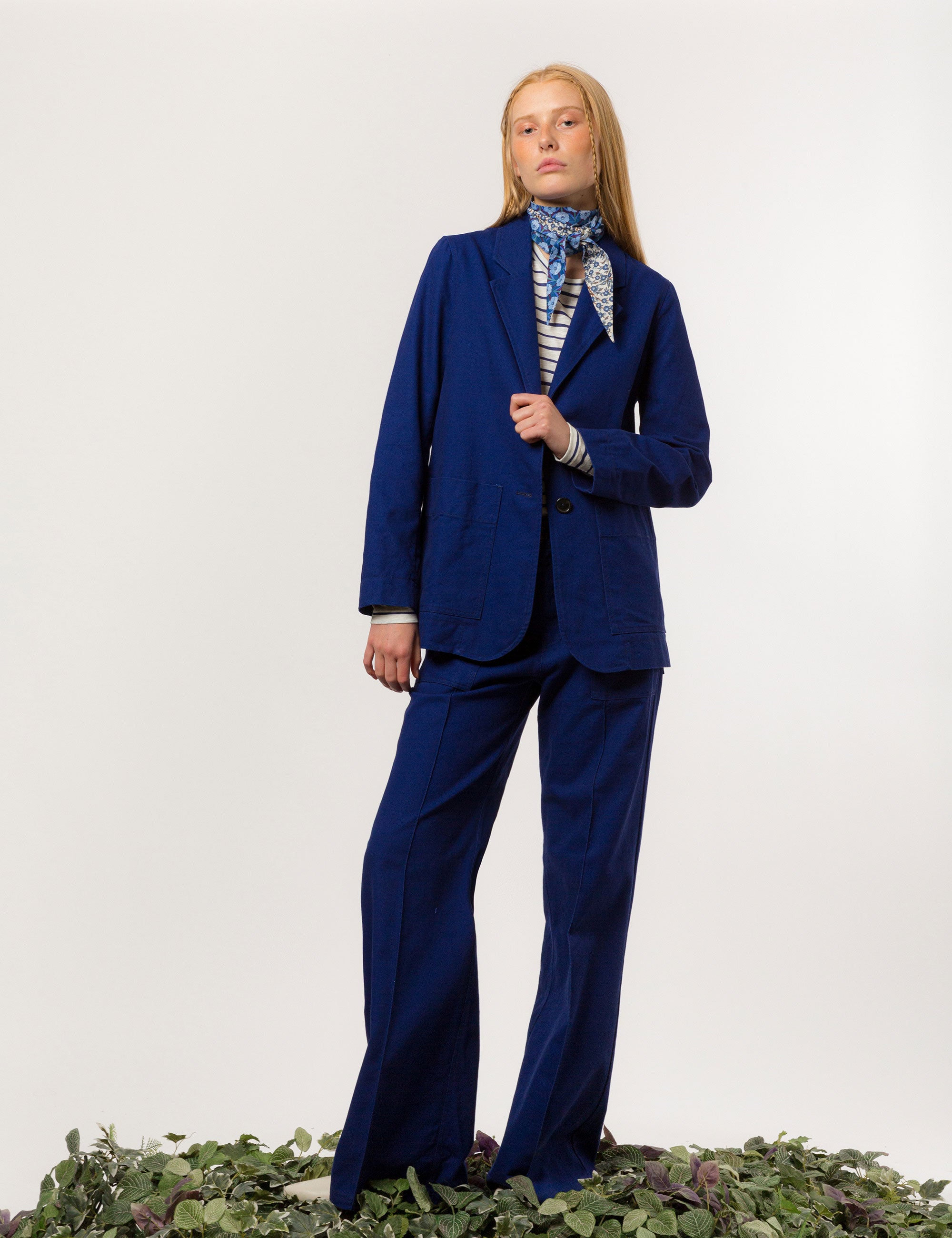 Woman in indigo blue canvas jacket blazer with buttons and pockets, matching wide canvas trousers, white and blue striped t-shirt and flower patterned bandana around neck