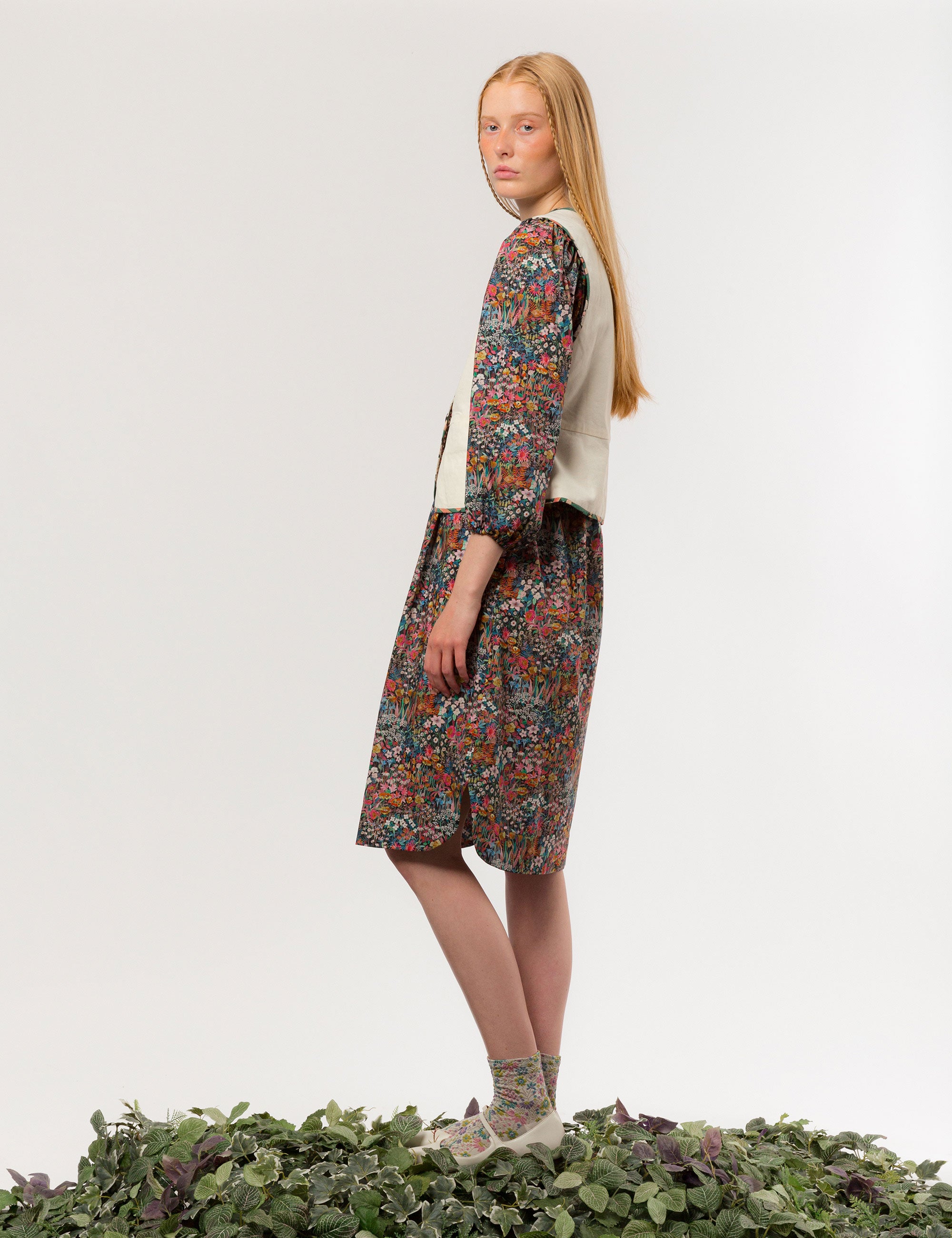 Woman in reversible cream colored vest with buttons, pockets and floral print pipings and multi-colored floral midi dress