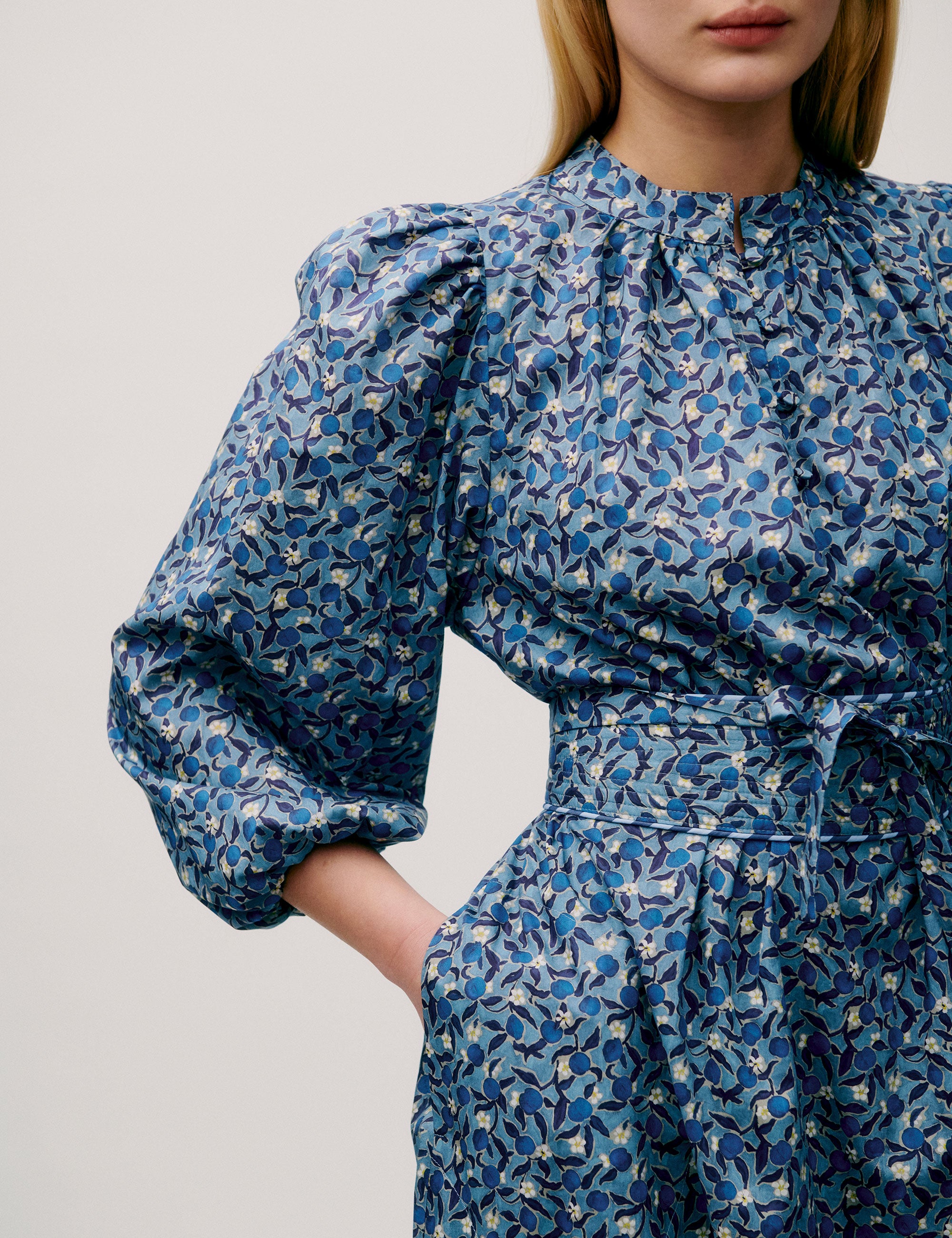 Woman wearing a long blue floral dress with puff sleeves, pleated neckline, and tie waist