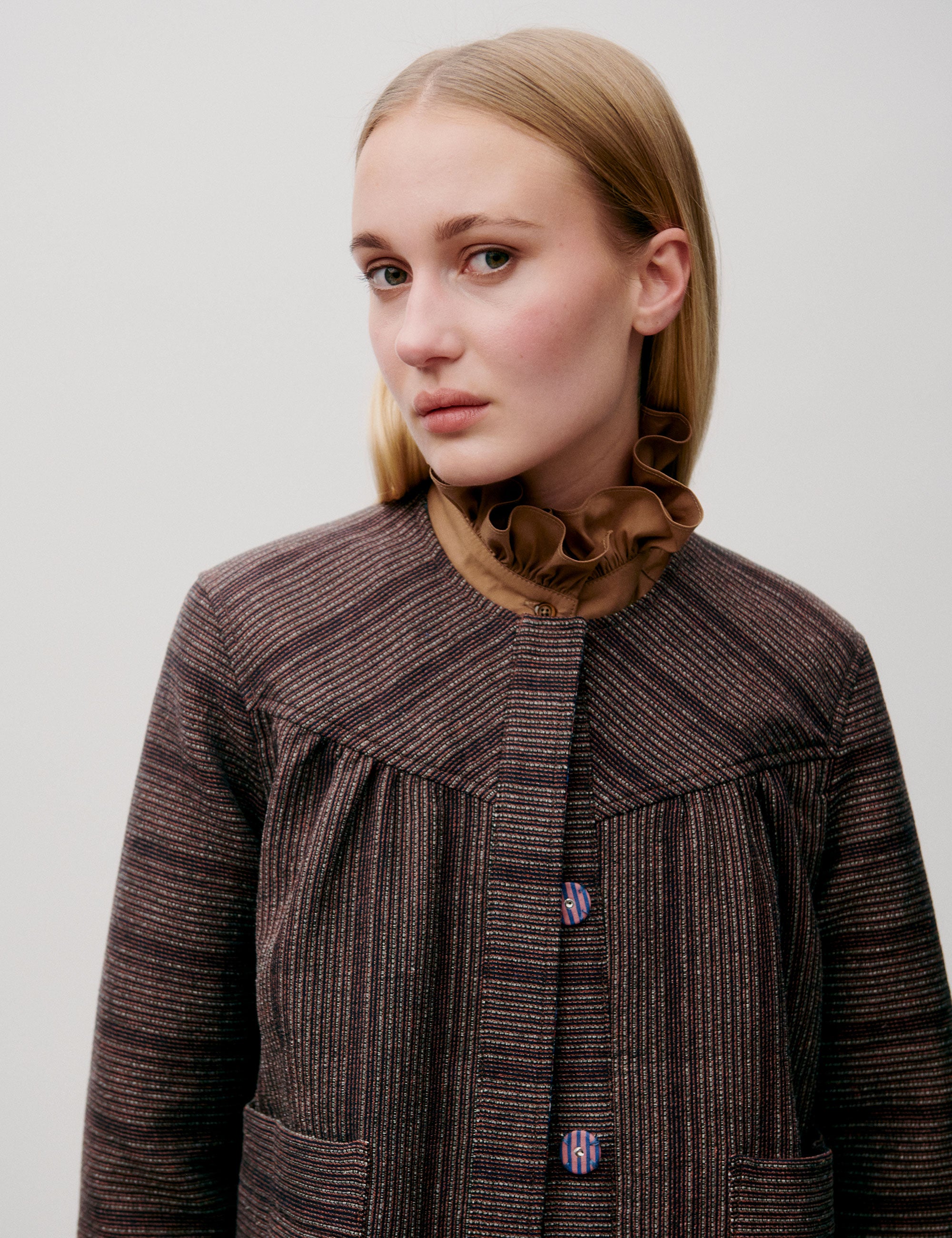 Woman in brown striped jacket with pockets and large buttons and brown shirt with frilled collar