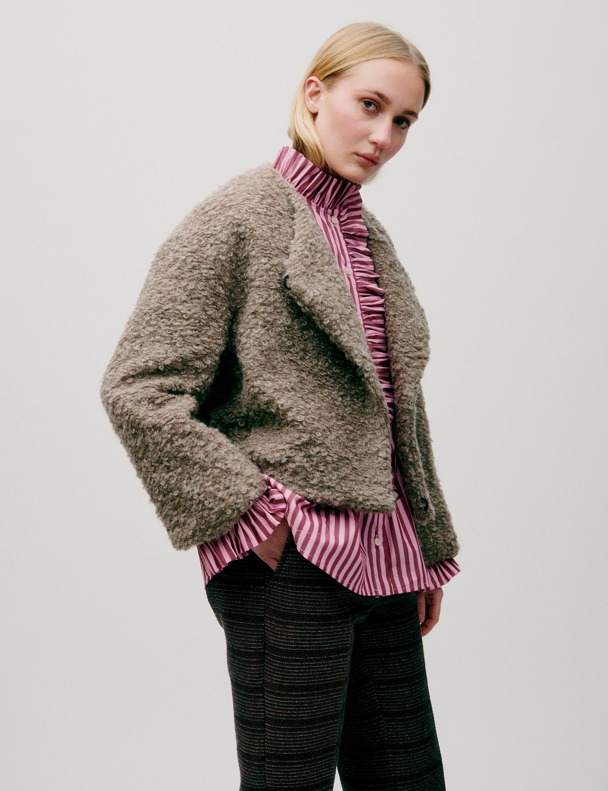 Woman in short wool jacket with assymetric design and a bubbly texture, a pink striped shirt with frilled chest and brown striped pants