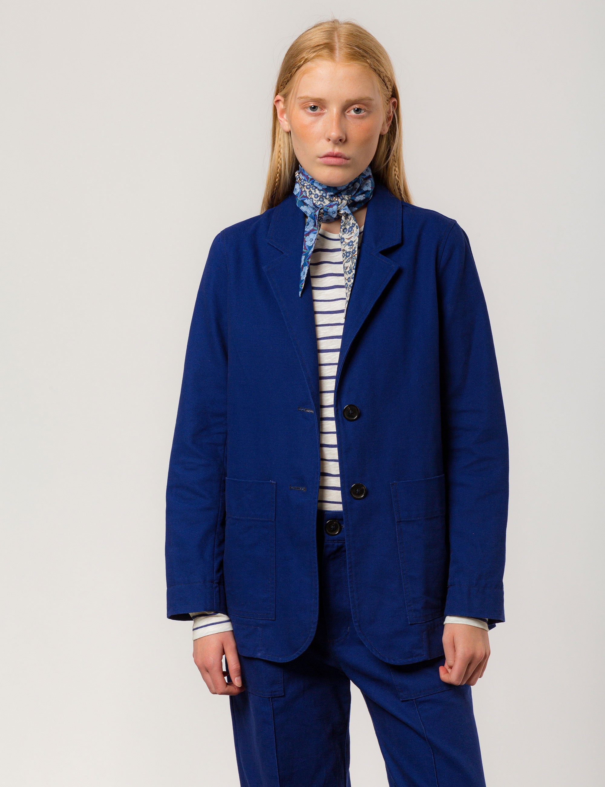 Woman in indigo blue canvas jacket blazer with buttons and pockets, matching wide canvas trousers, white and blue striped t-shirt and flower patterned bandana around neck