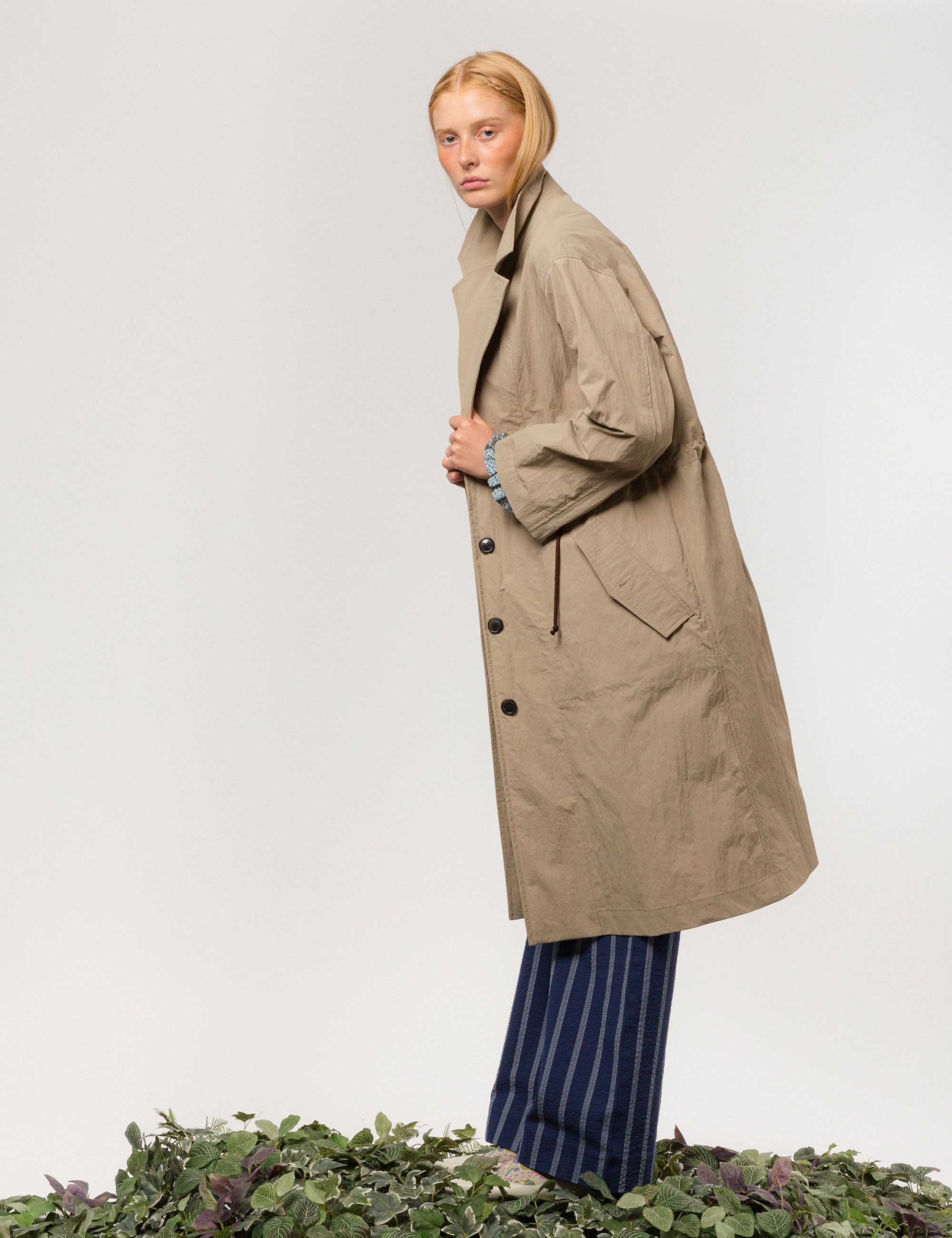 Women in long khaki coat with buttons, large pockets and floral print pipings, wearing blue and white striped pants