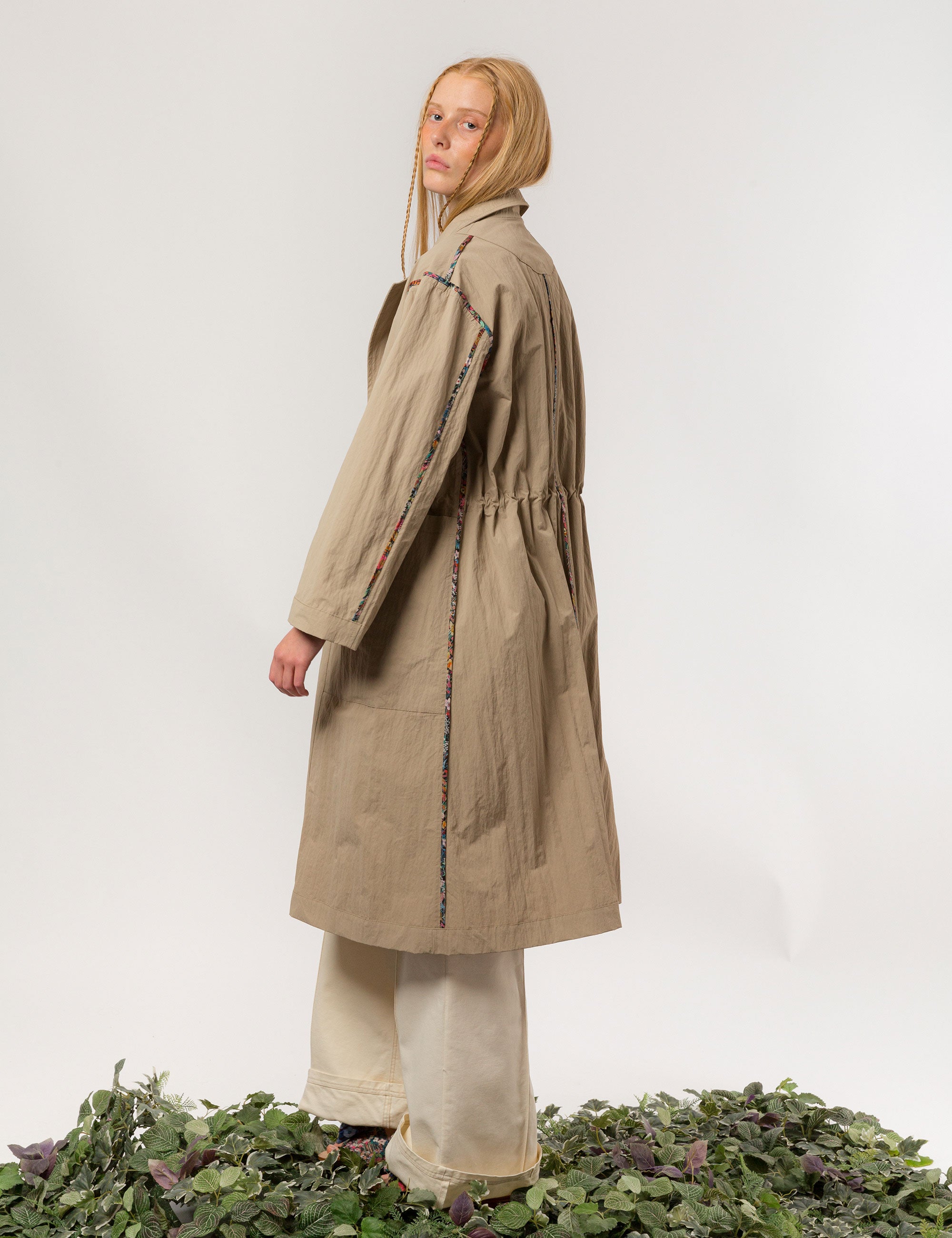 Women in long khaki coat with buttons, large pockets and floral print pipings, and white trousers