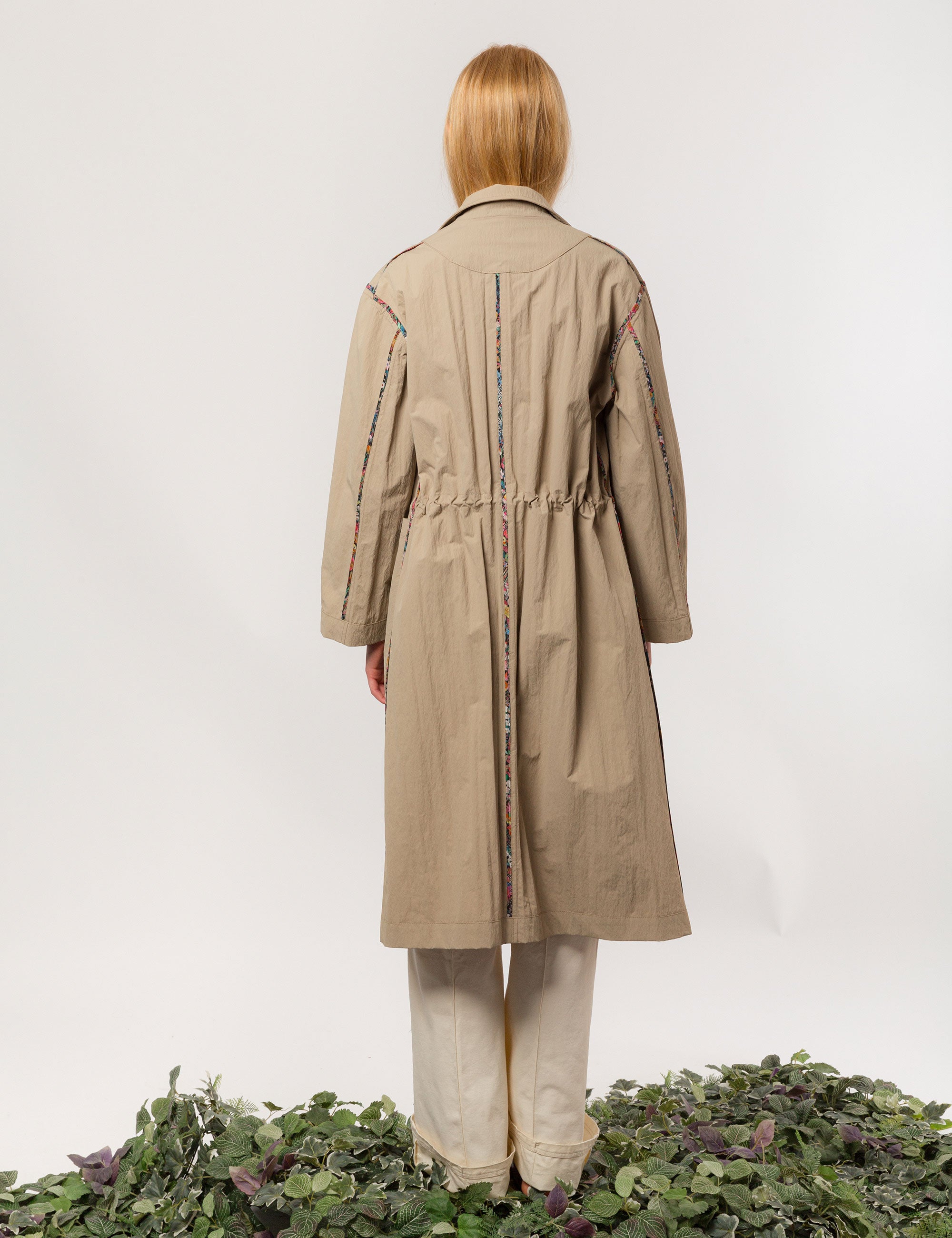 Women in long khaki coat with buttons, large pockets and floral print pipings, wearing white trousers