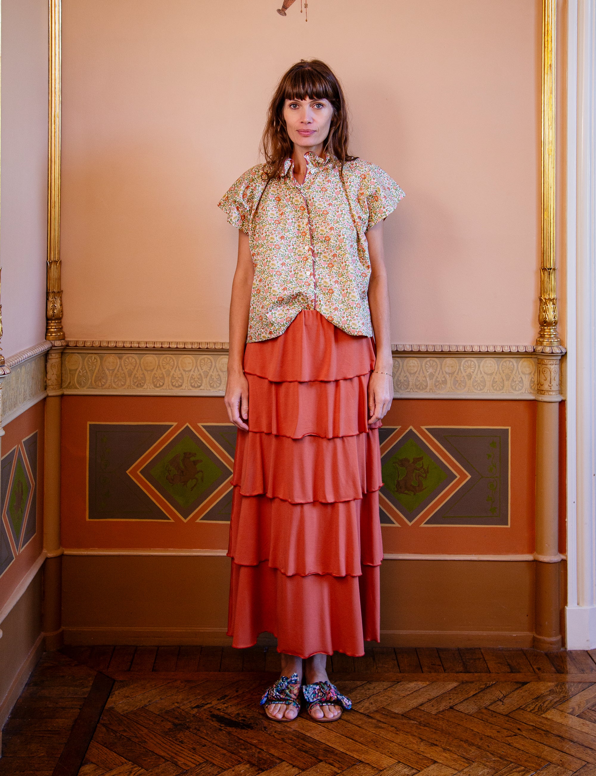 Woman wearing a floral blouse and tiered orange skirt in an ornate room.