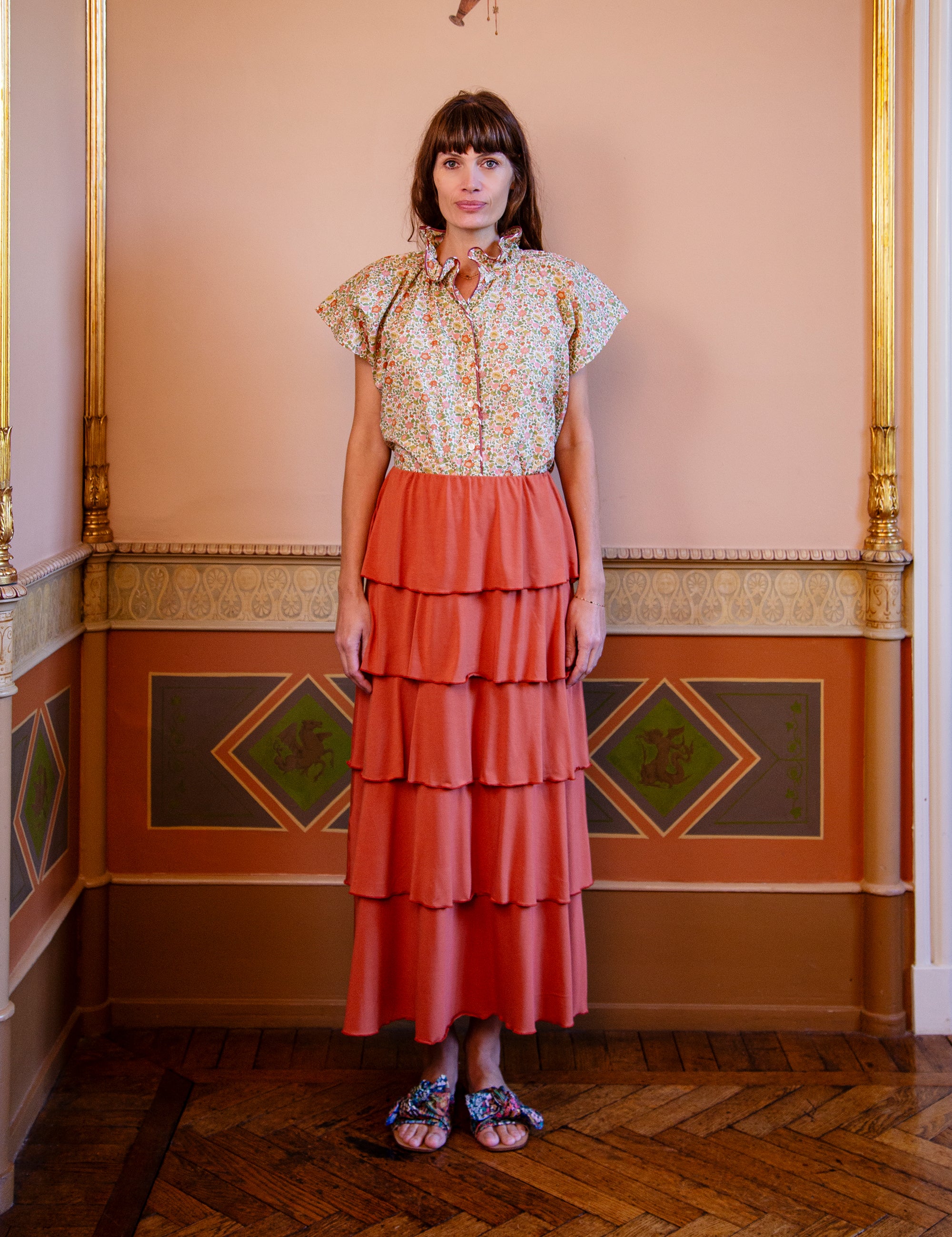 Woman wearing a floral blouse and tiered coral skirt in an ornate room.