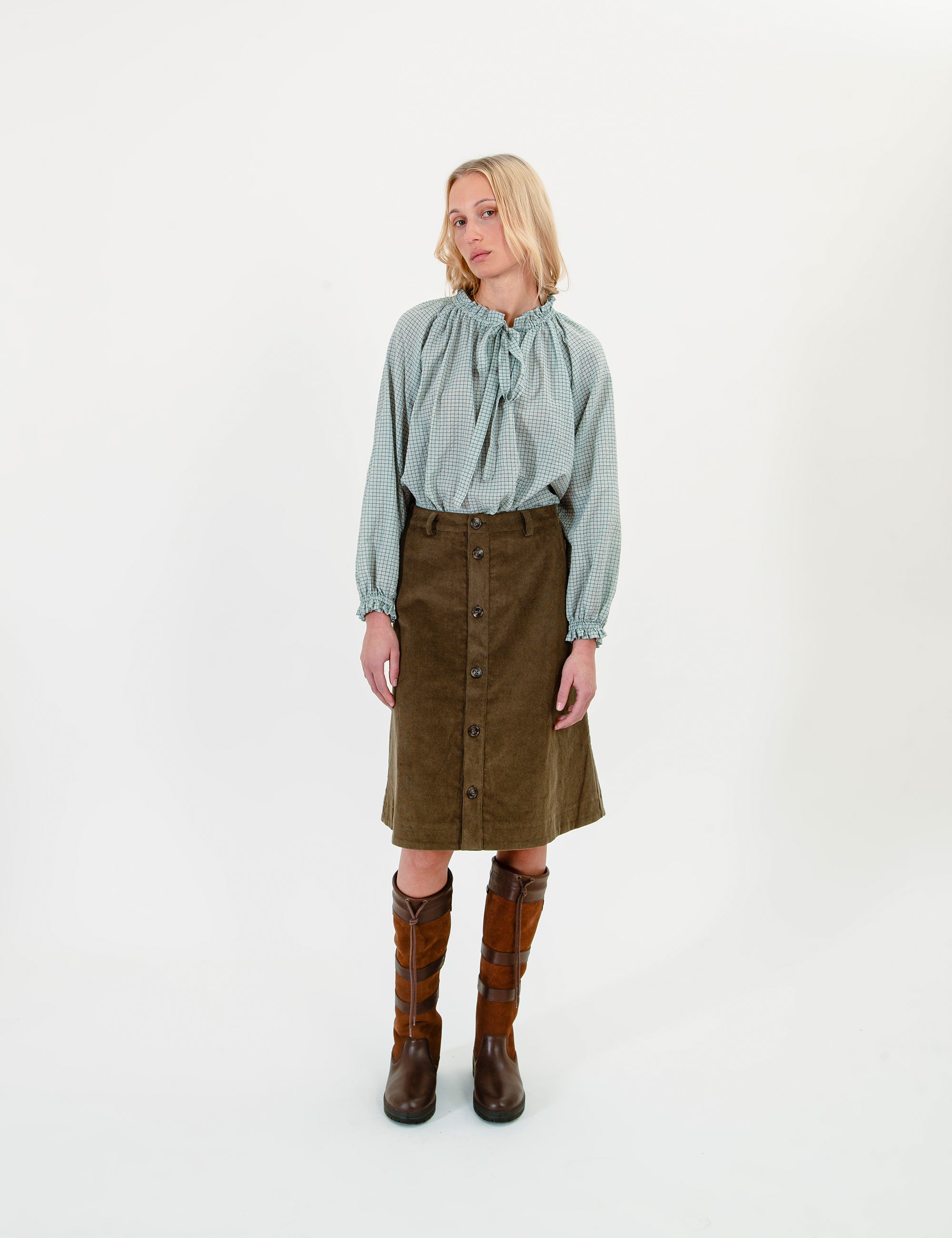 Woman wearing a light green blouse, olive green corduroy skirt, and brown boots on a white background