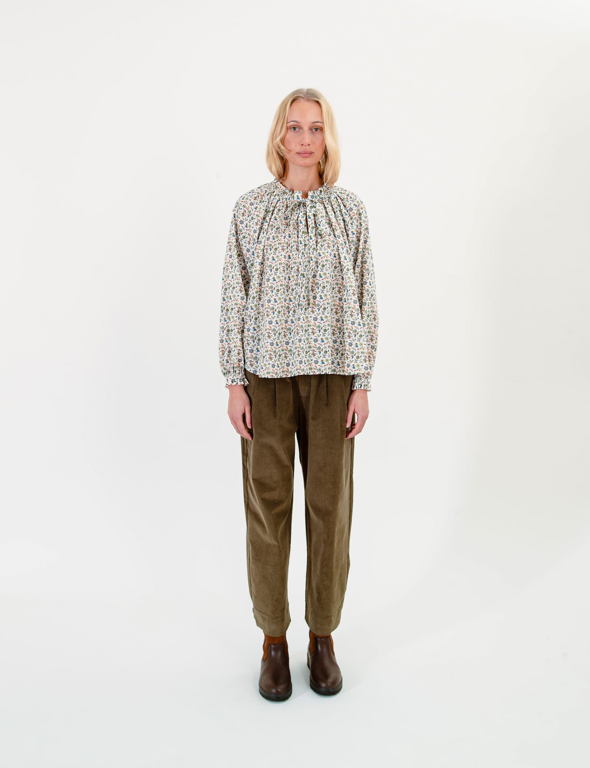 Woman wearing a patterned blouse and olive green corduroy pants on a white background
