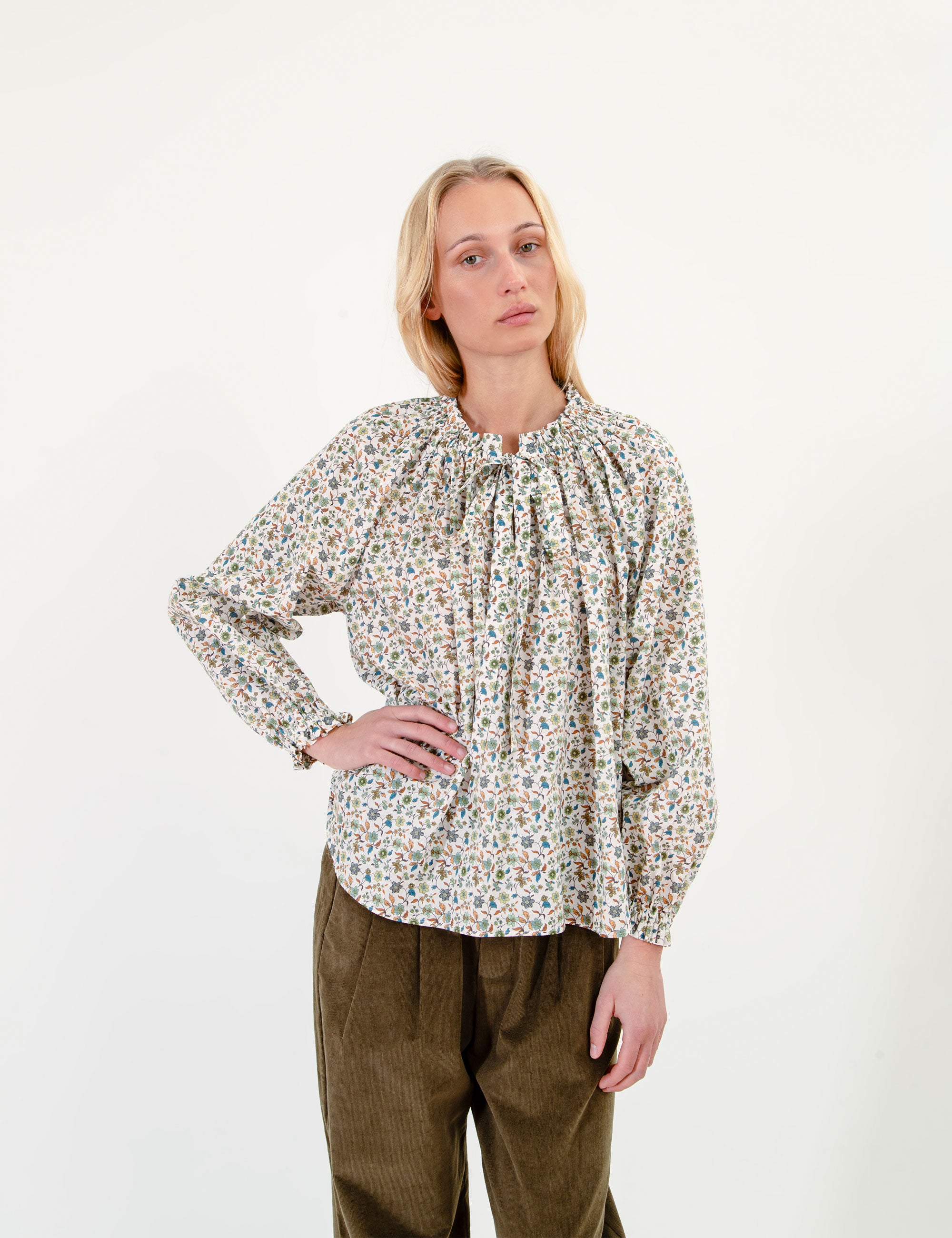 Woman wearing a patterned blouse and olive green corduroy pants on a white background