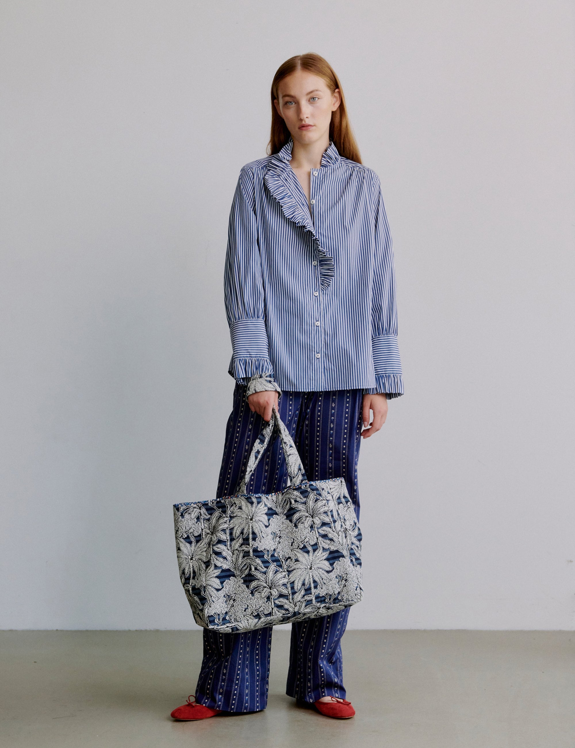 Woman holding a blue floral tote bag wearing a blue striped shirt and blue patterned pants on a plain background