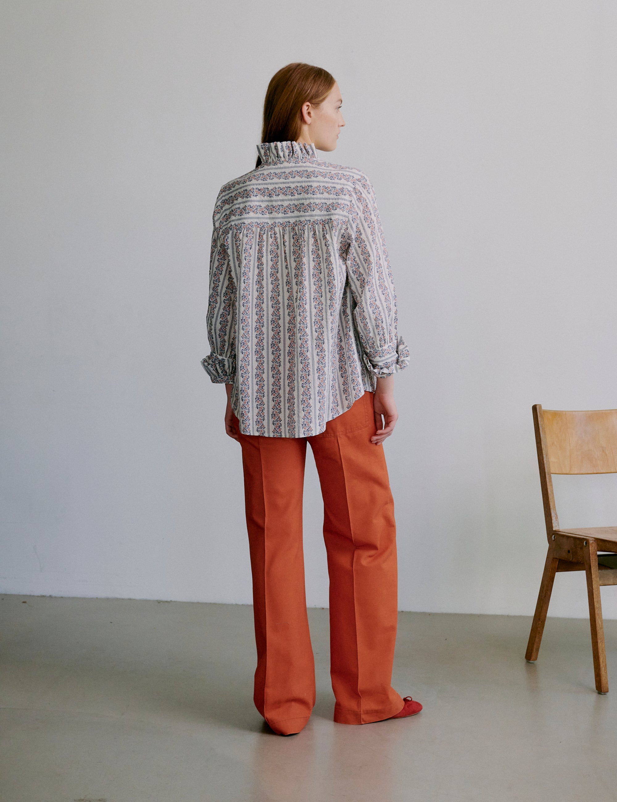 Woman wearing a floral patterned blouse with frills and orange pants standing against a plain background with a wooden chair.