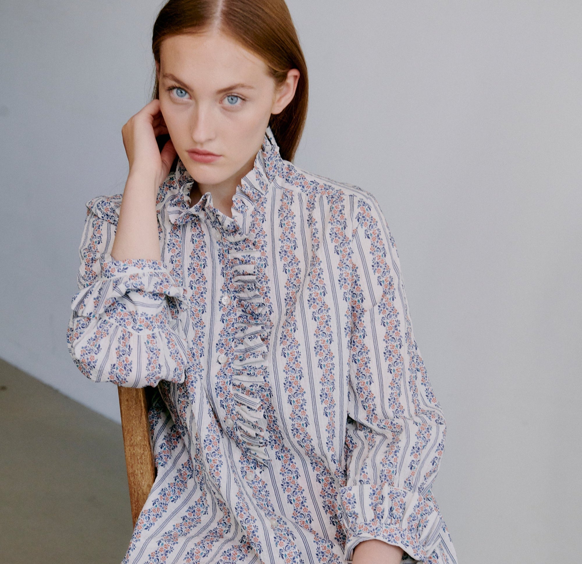 Woman wearing a patterned blouse and orange pants sitting on a wooden chair.
