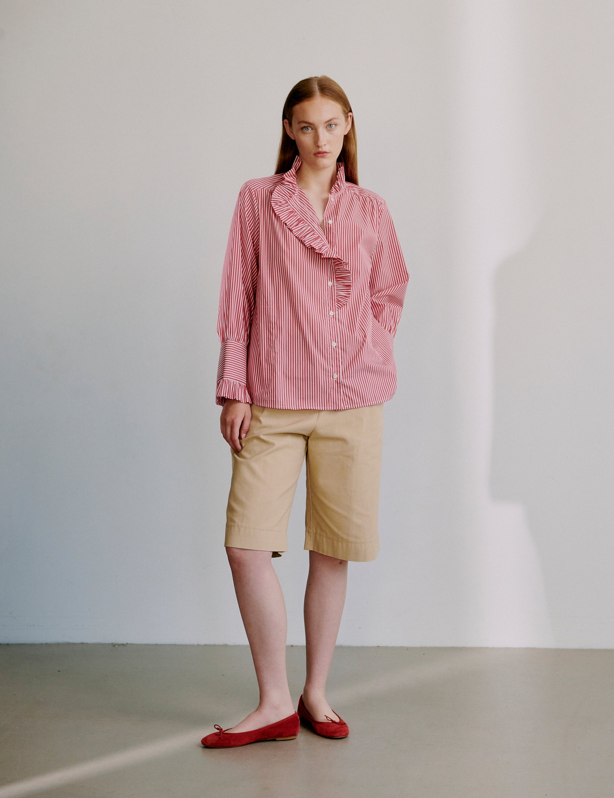 Woman wearing a pink striped shirt with frilled details and beige shorts against a plain background