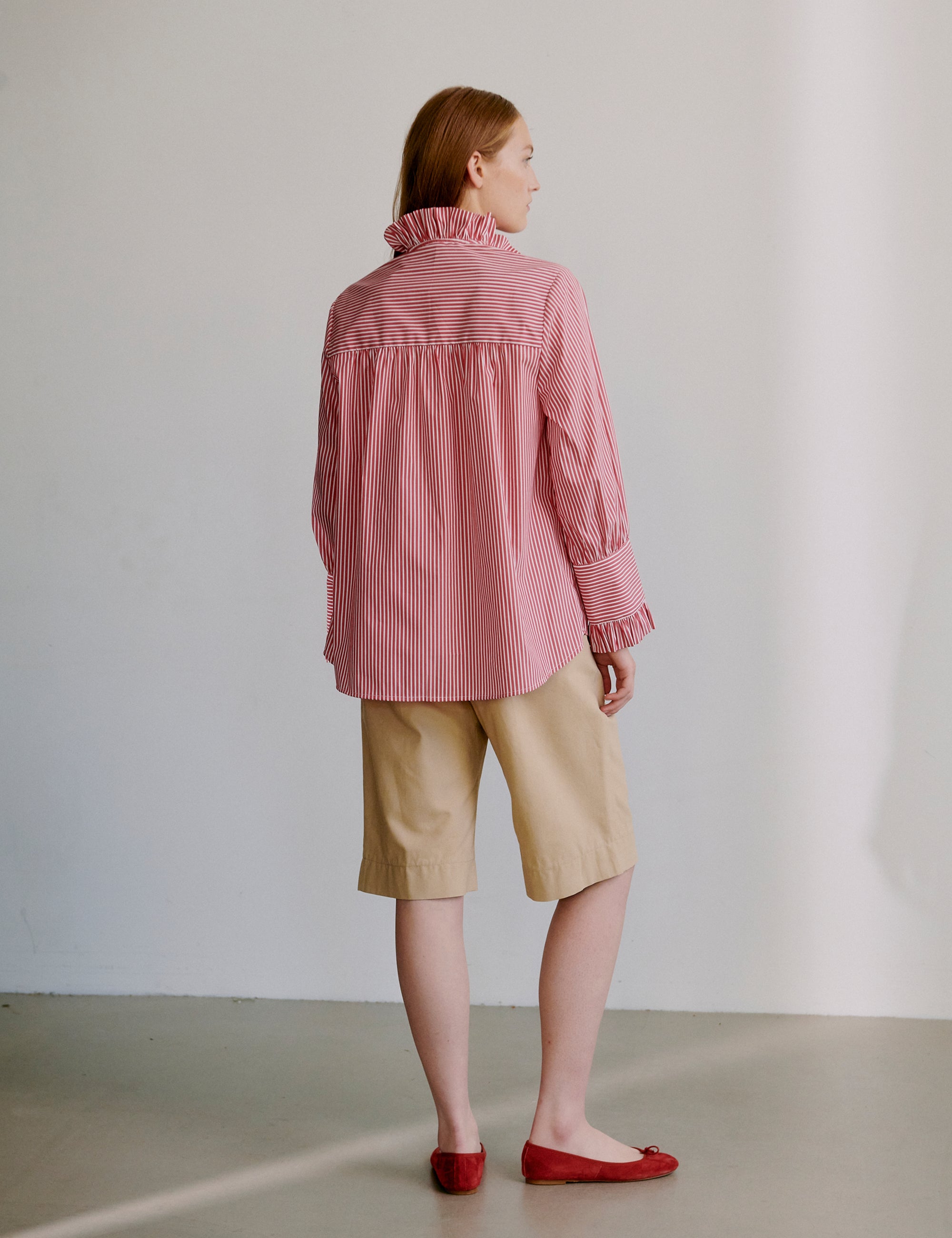 Woman wearing a pink striped shirt with frilled details and beige shorts against a plain background