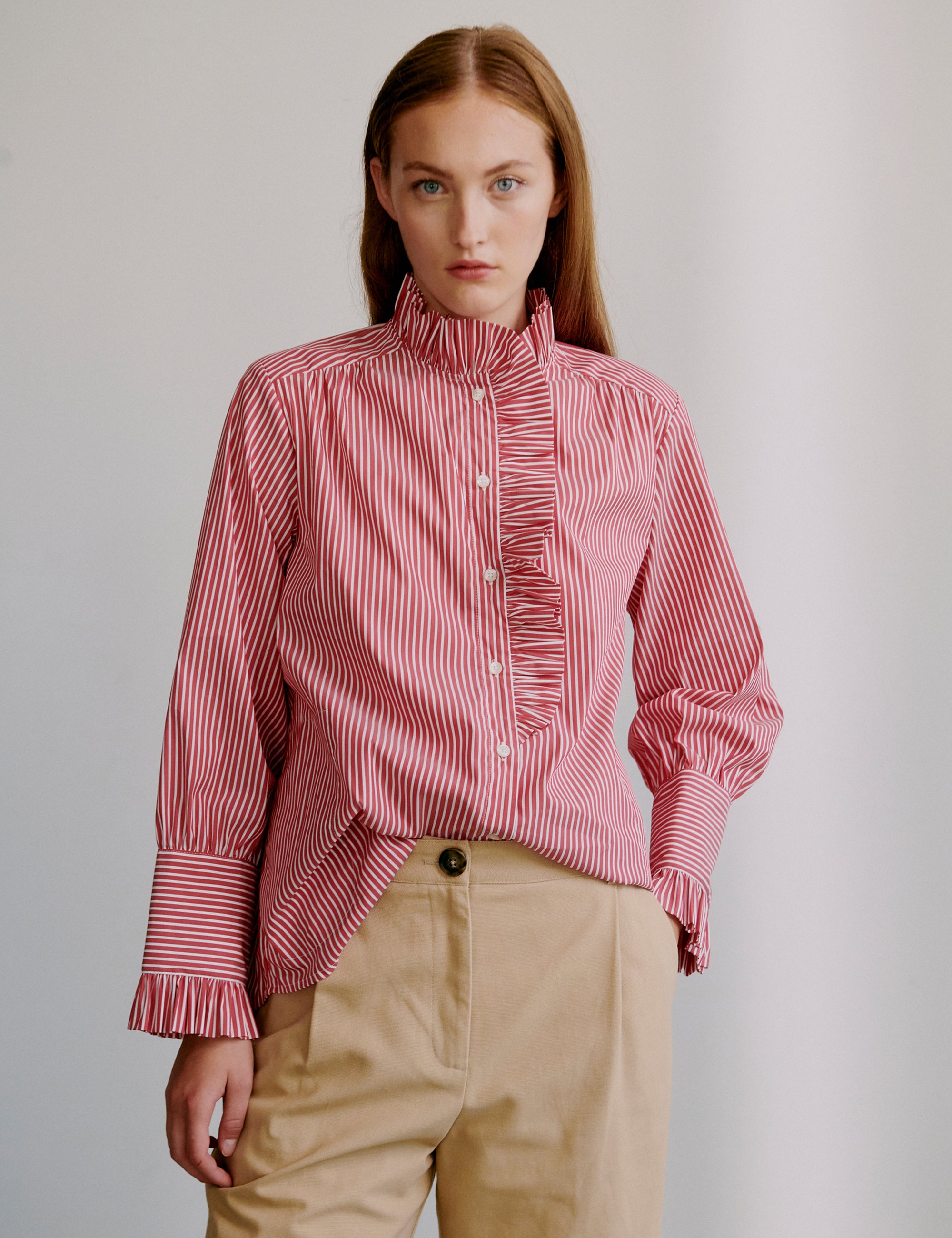 Woman wearing a pink striped shirt with frilled details and beige shorts against a plain background