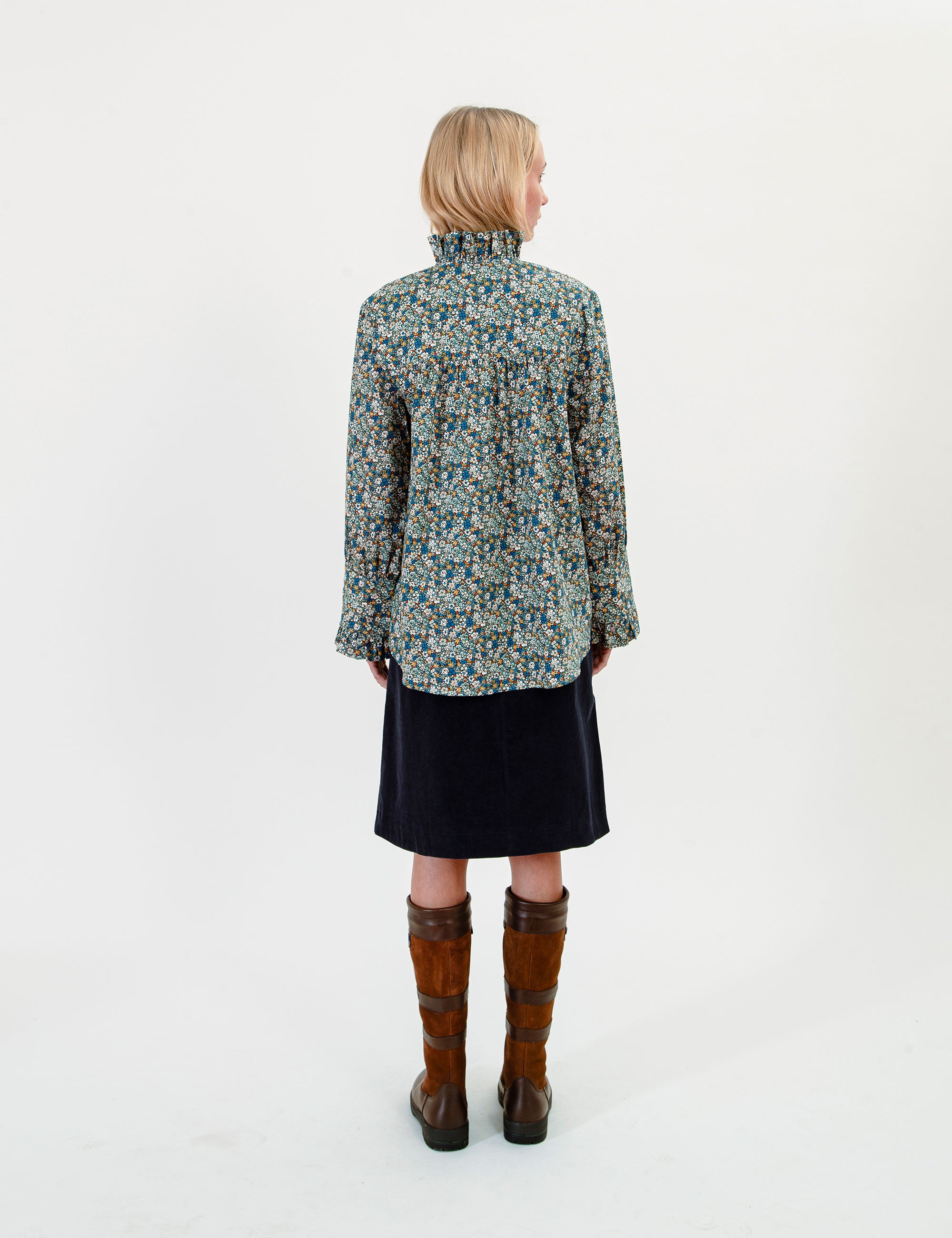 Woman wearing a floral blouse with ruffled stand collar, dark corduroy skirt, and brown boots on a white background 