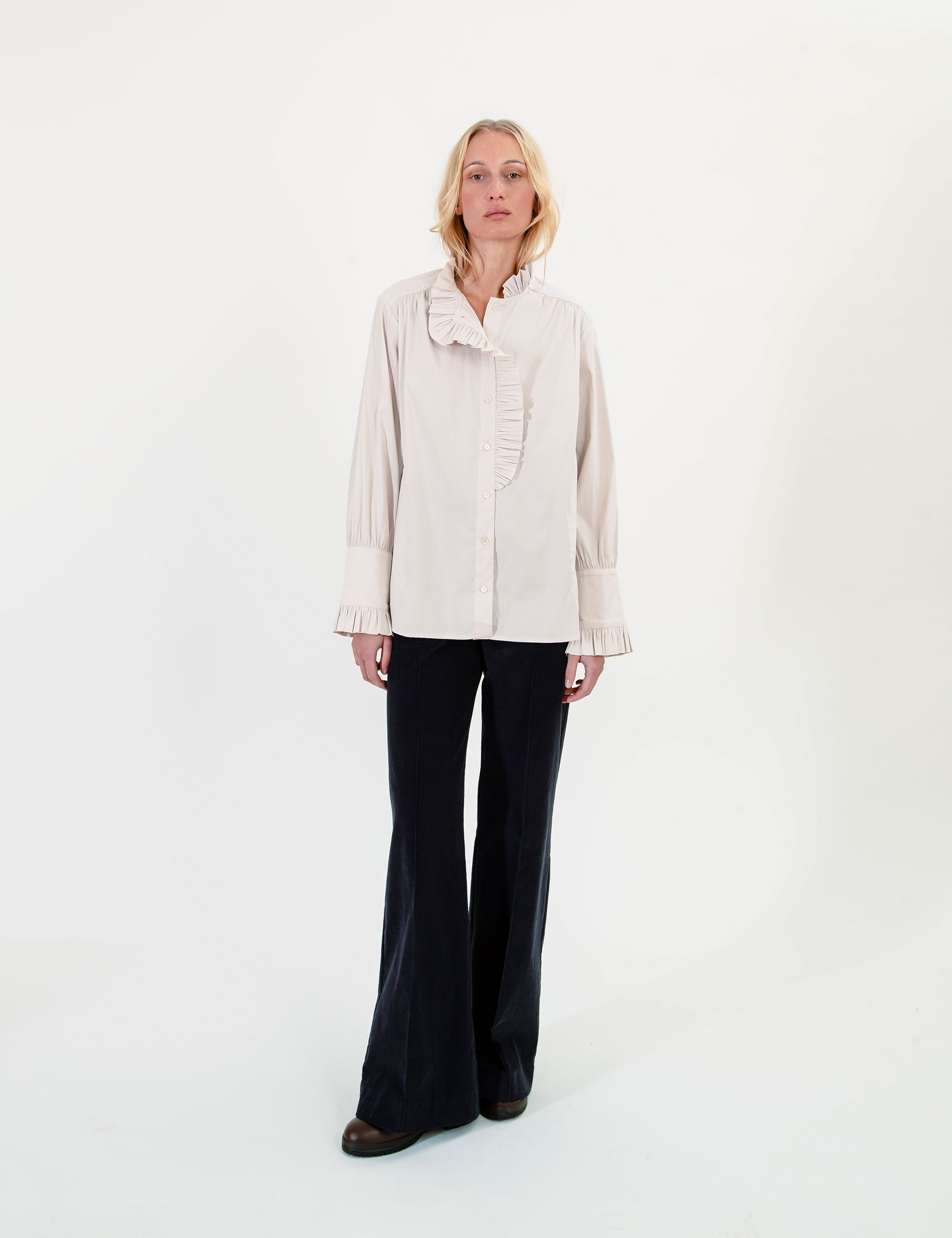 Woman wearing a light-colored ruffled blouse and dark pants on a white background
