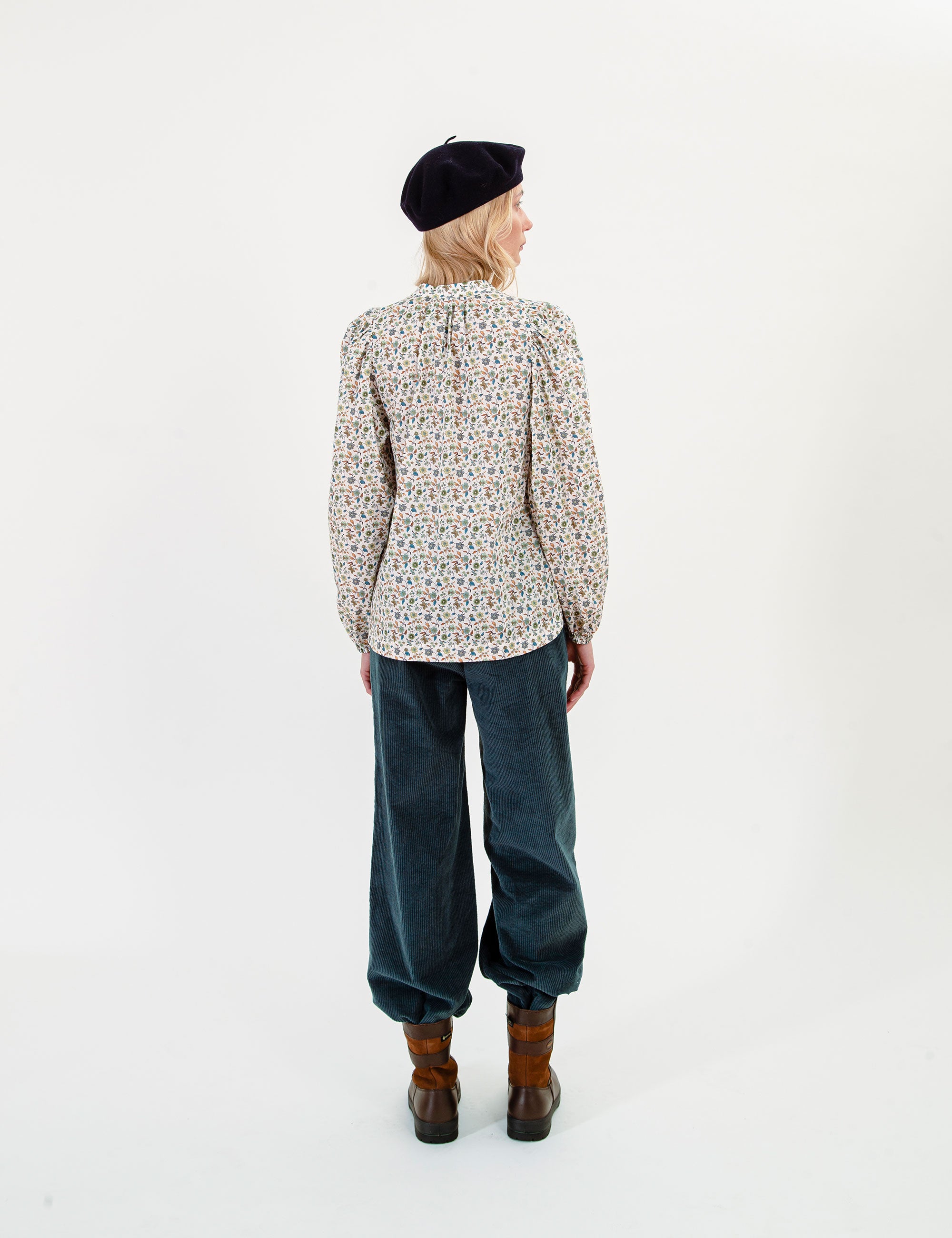 Woman wearing a floral patterned blouse, dark blue corduroy pants, and a beret on a white background