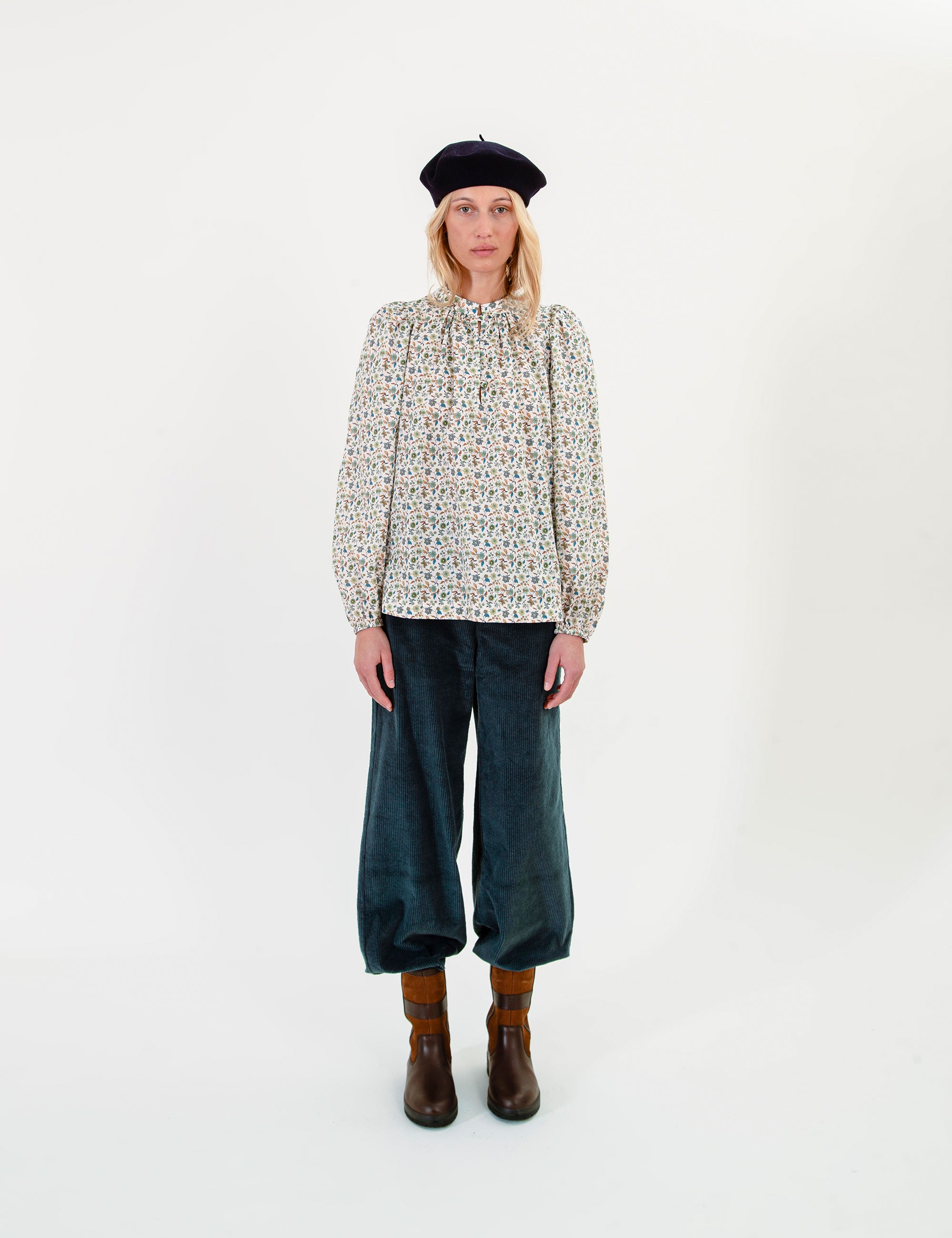 Woman wearing a floral patterned blouse, dark pants, and a beret on a white background