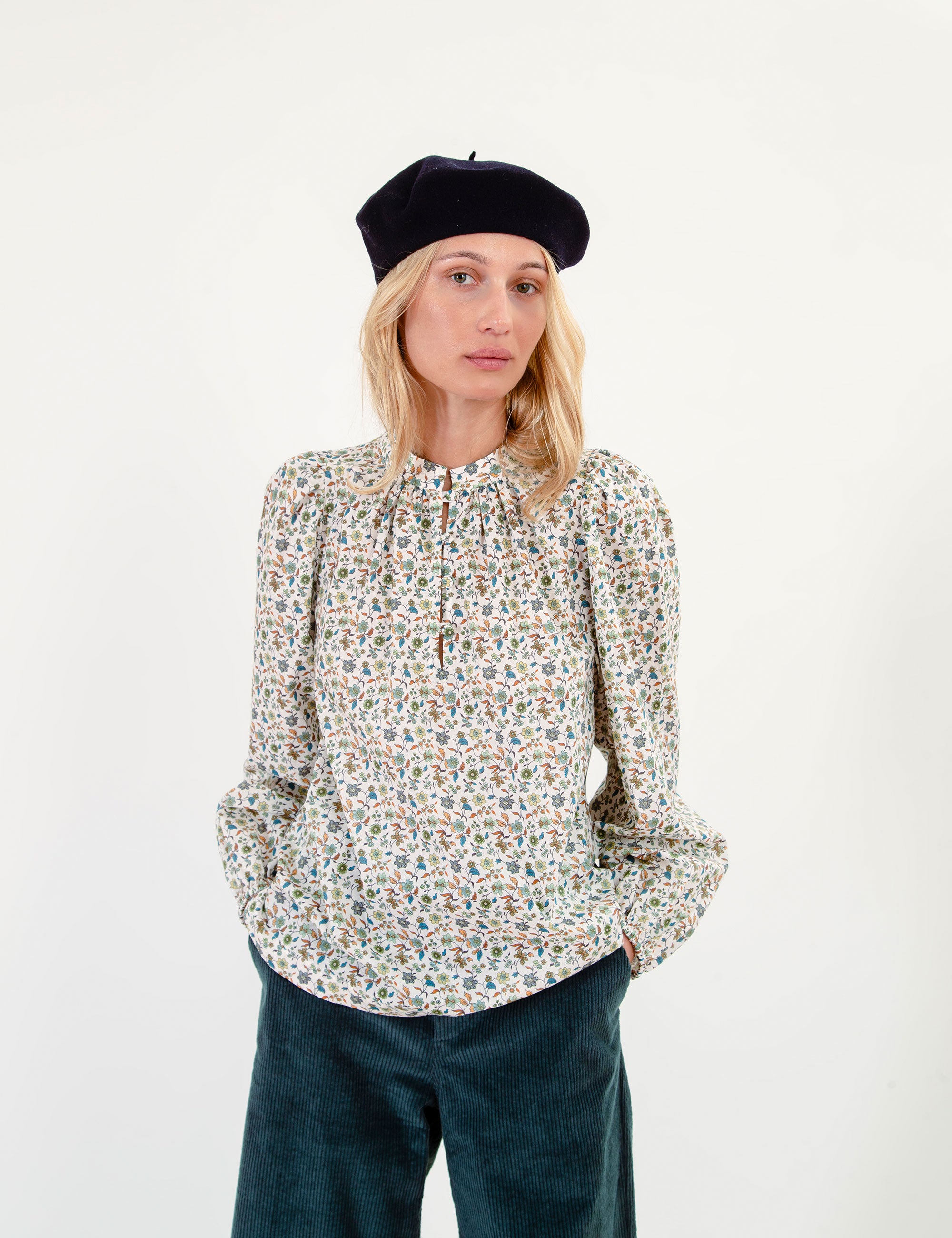 Woman wearing a floral patterned blouse, dark blue corduroy pants, and a beret on a white background