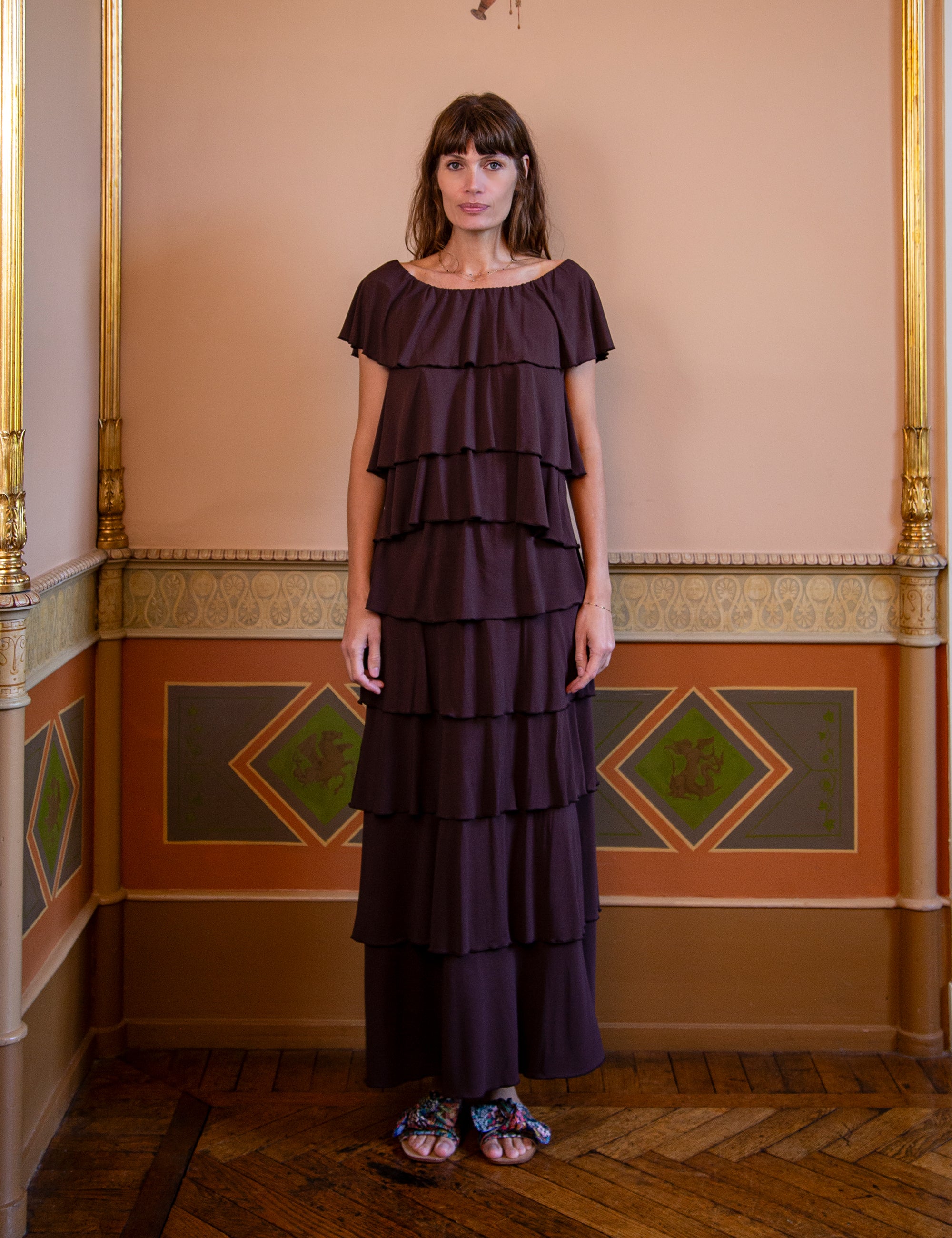 Woman wearing a dark brown tiered skirt and matching top standing in an ornate room with decorative walls.