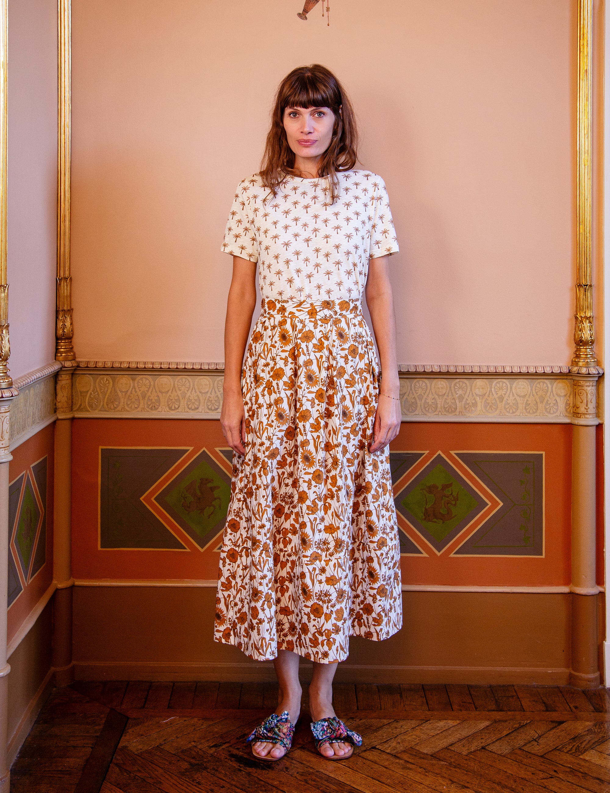Woman wearing a white t-shirt with palm print and floral patterned skirt standing in an ornate room.