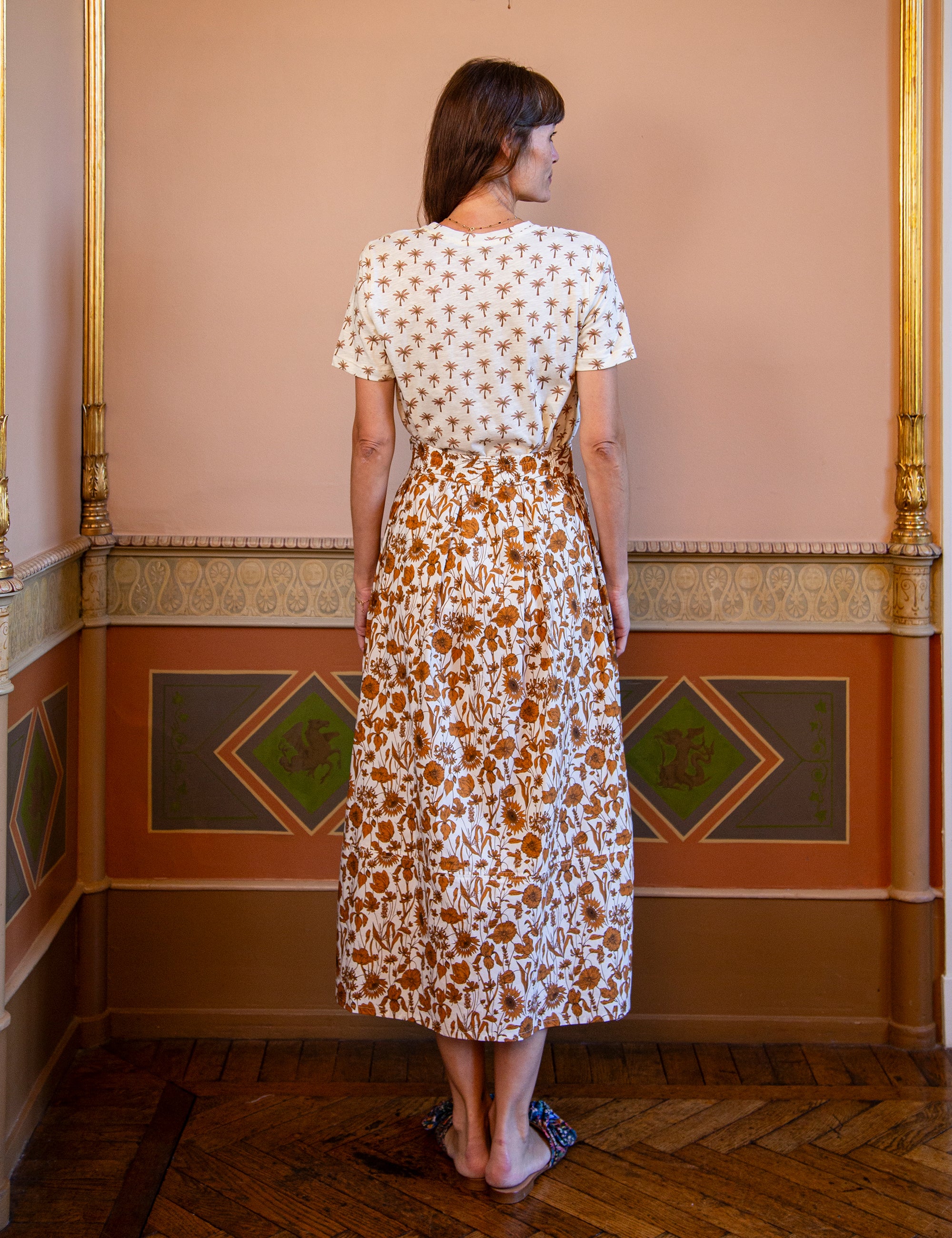 Woman wearing a white t-shirt with palm print and floral patterned skirt standing in an ornate room.
