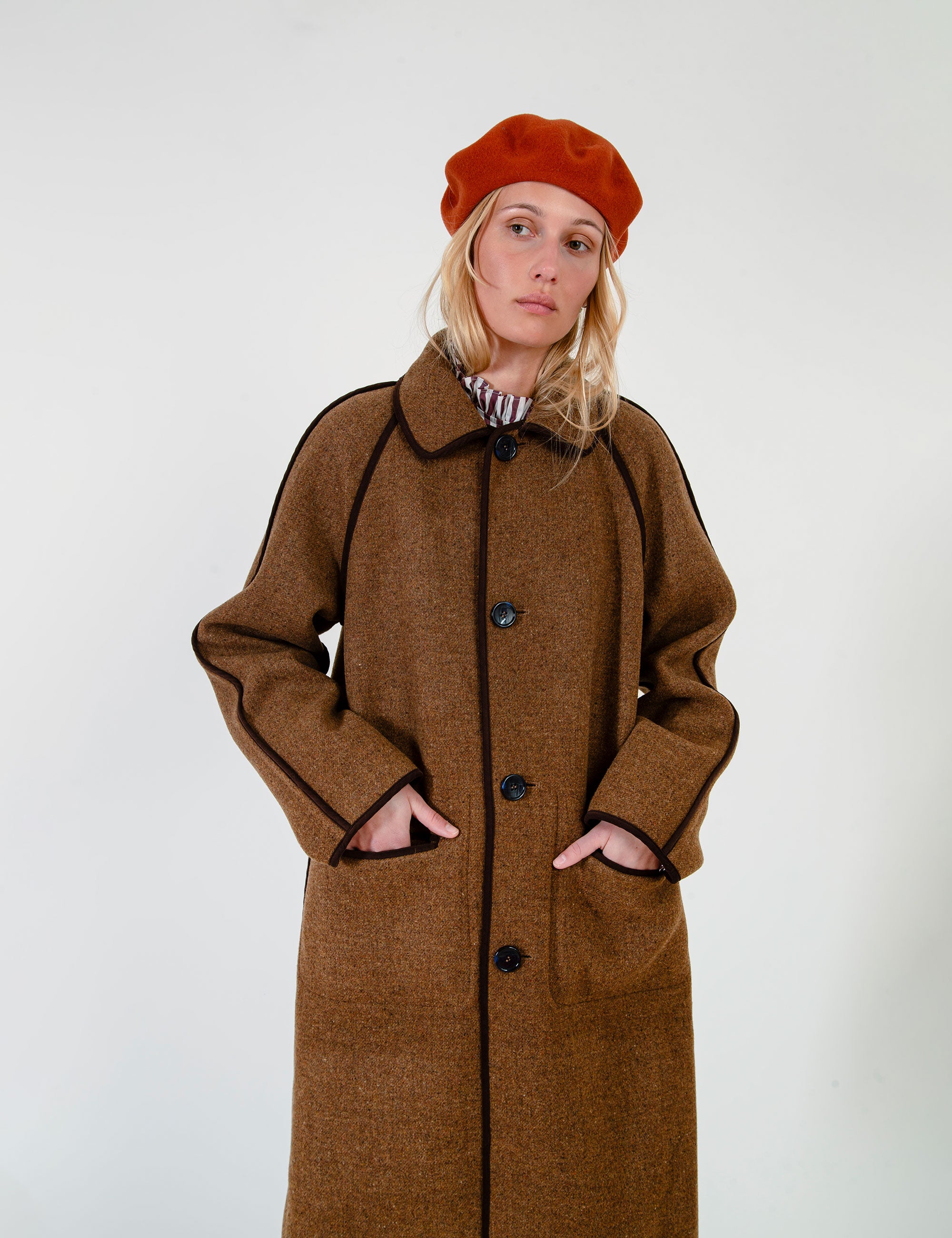Woman wearing a long brown coat with pockets and collar and a red beret on a white background