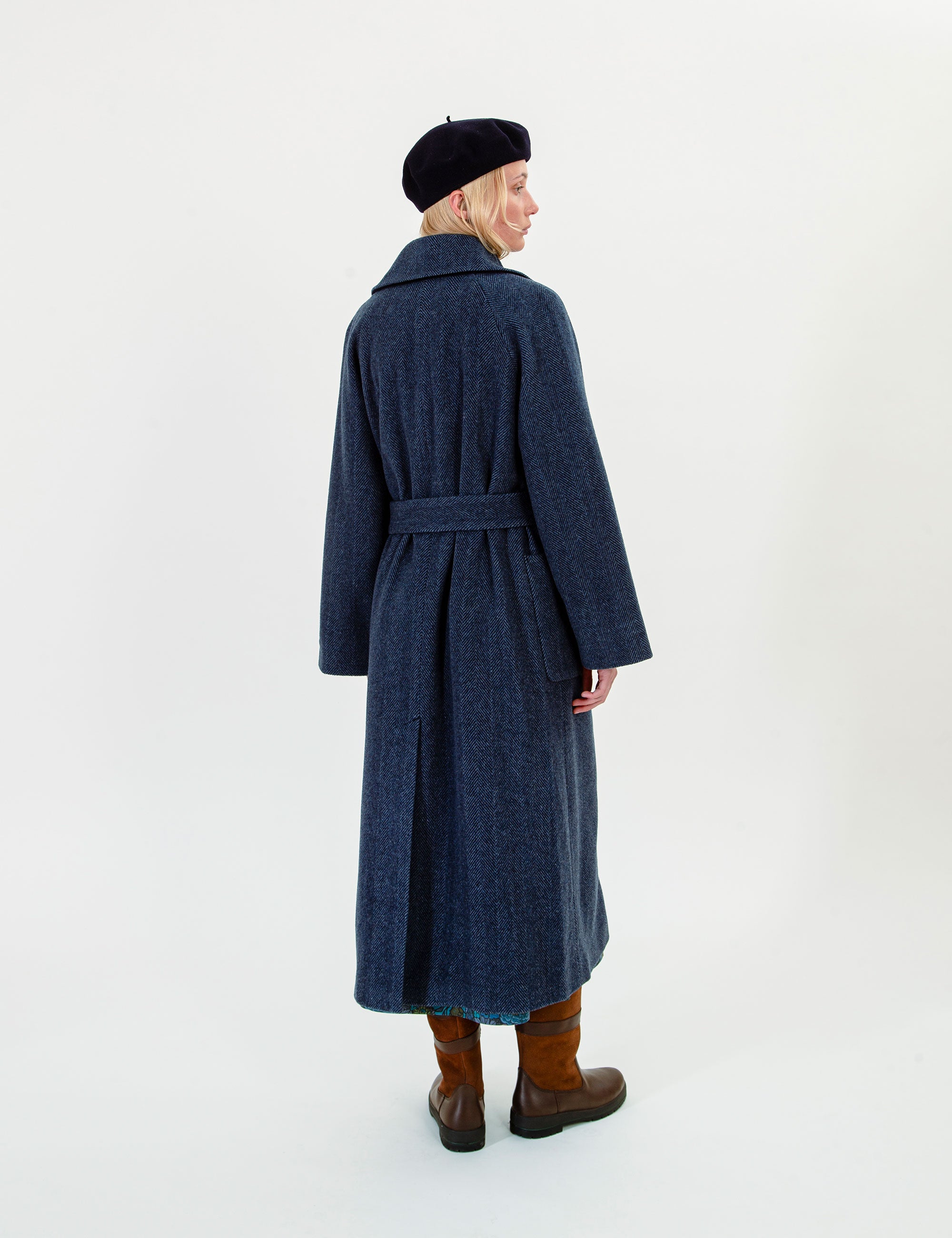 Woman in a long navy blue wool coat with button details, belt and pockets on a light gray background
