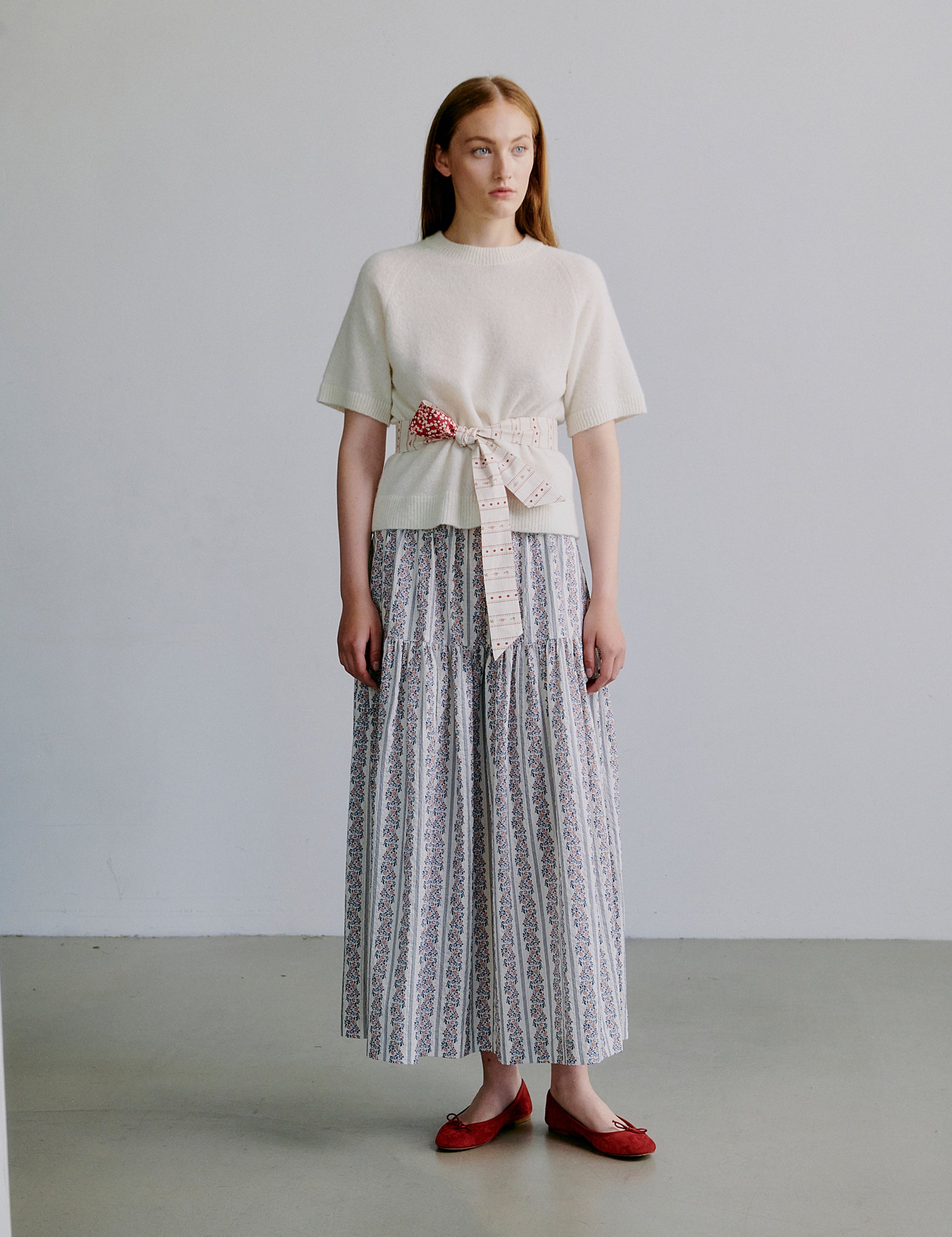 Woman wearing a light-colored knit top and floral patterned skirt with fabric belt and red shoes against a plain background