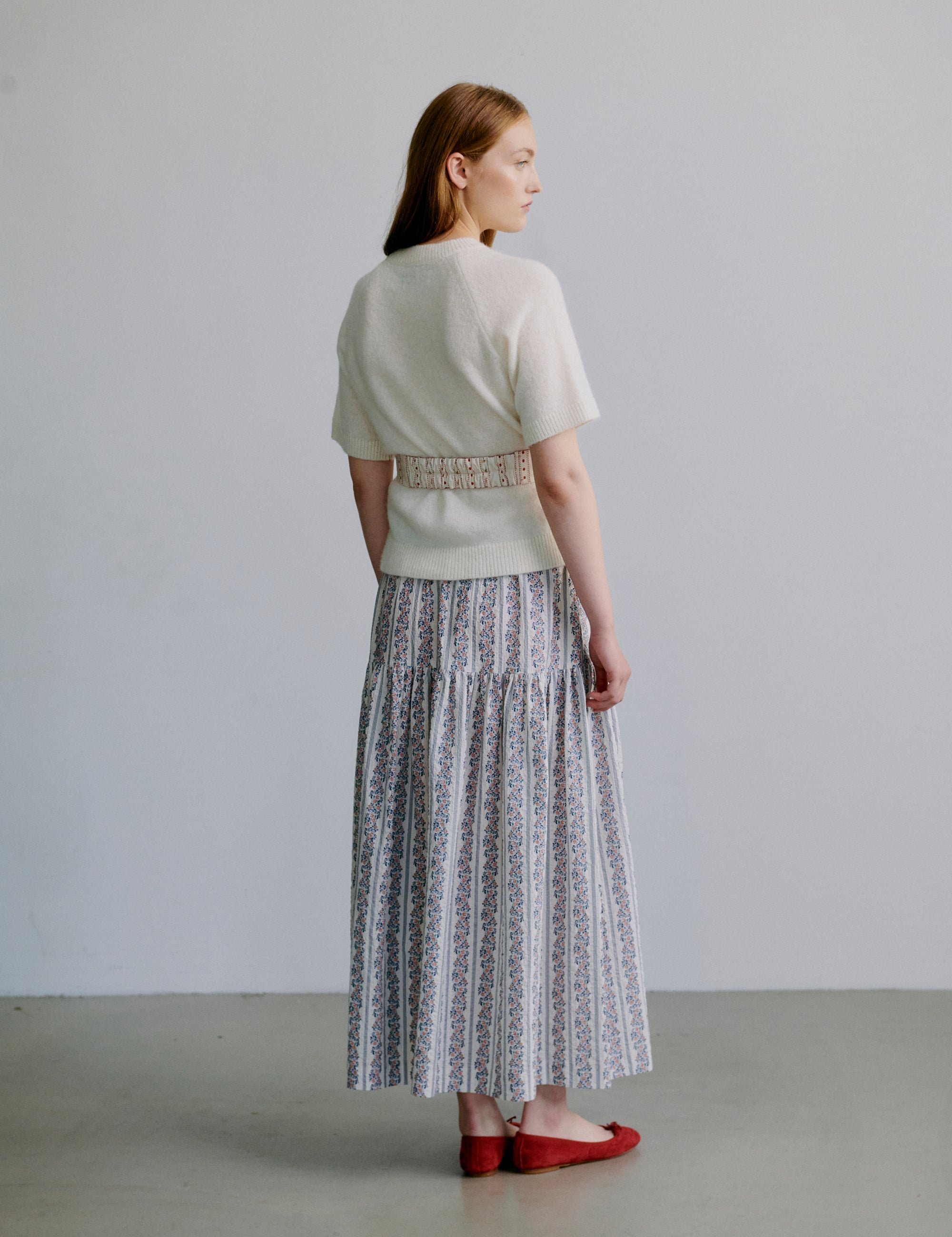 Woman wearing a white short sleeved knit  top and floral patterned skirt with red shoes against a plain background