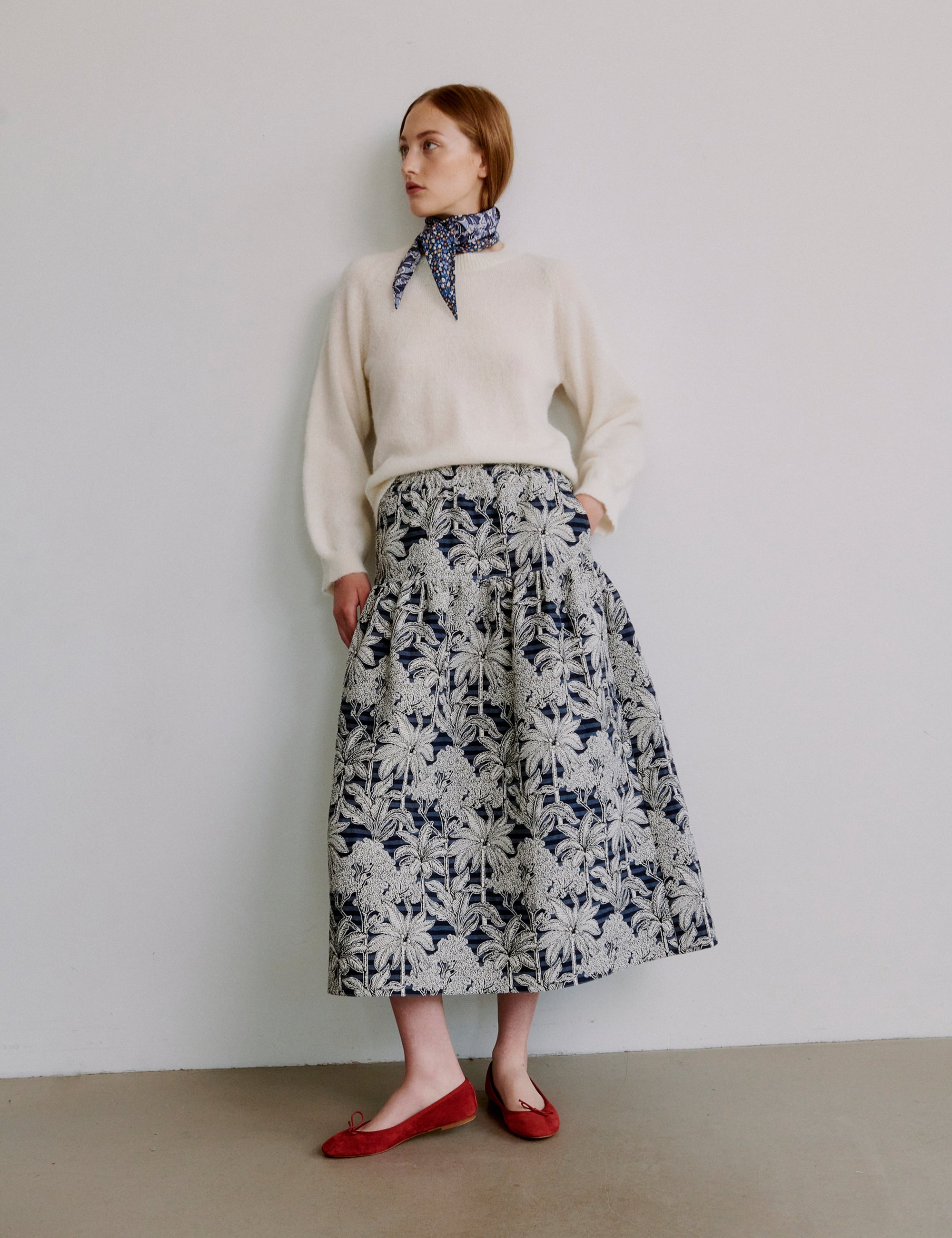 Kvinde iført en cremefarvet sweater og blomstret nederdel mod hvid baggrund