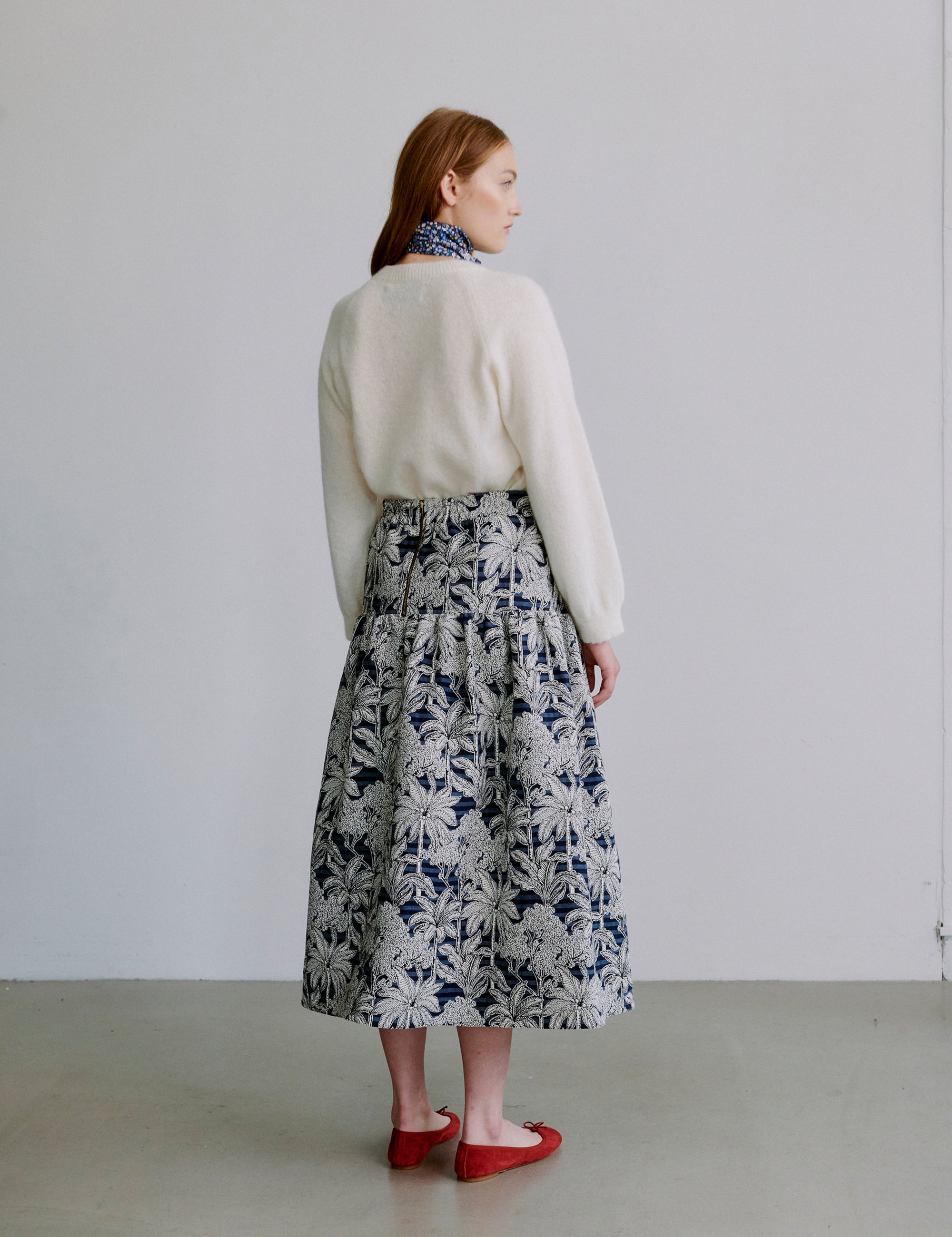 Woman wearing a white sweater and floral skirt with red shoes against a plain background