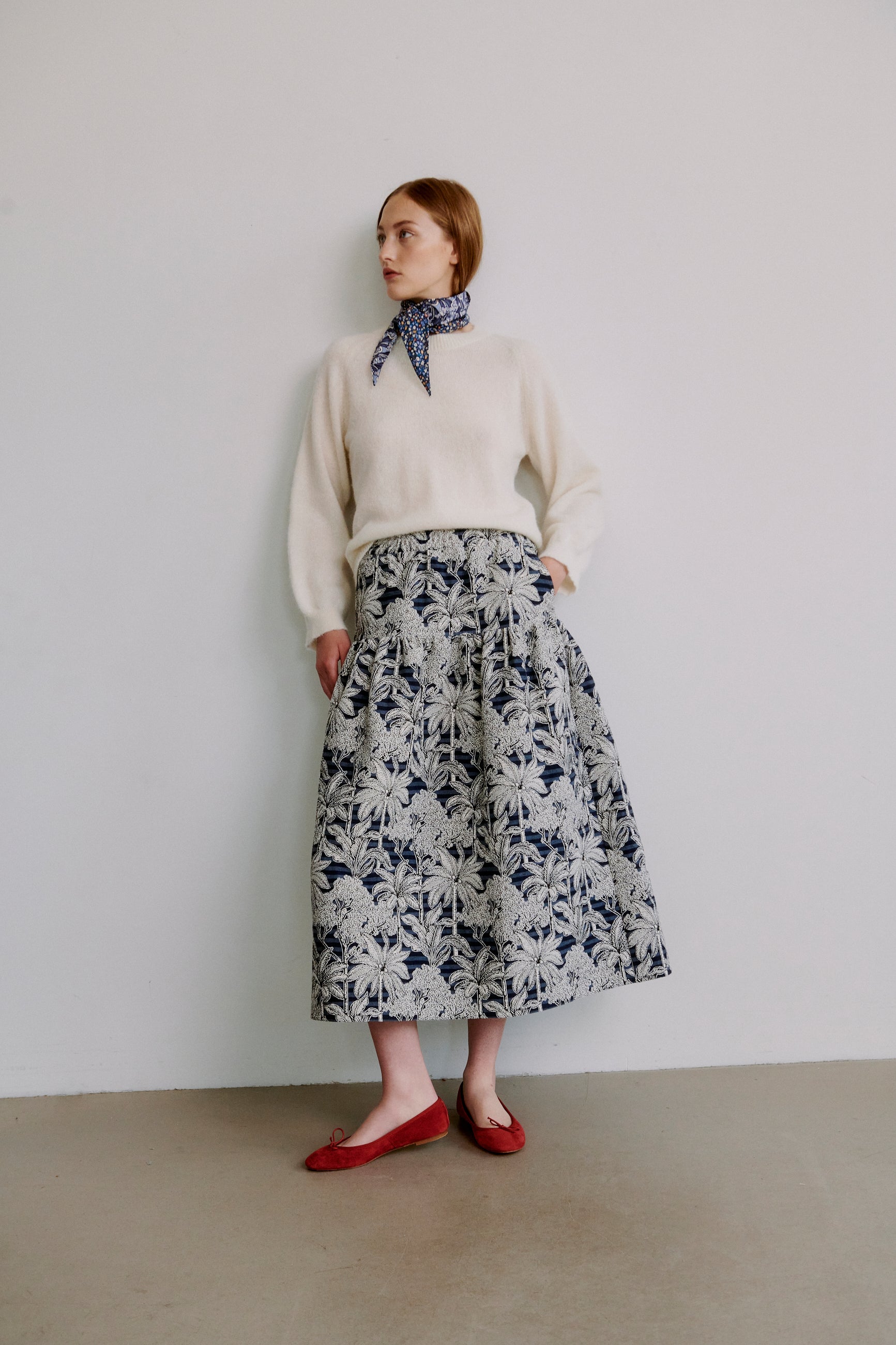 Woman wearing a cream colored long sleevew knit sweater and blue floral skirt with a blue patterned scarf against a plain background