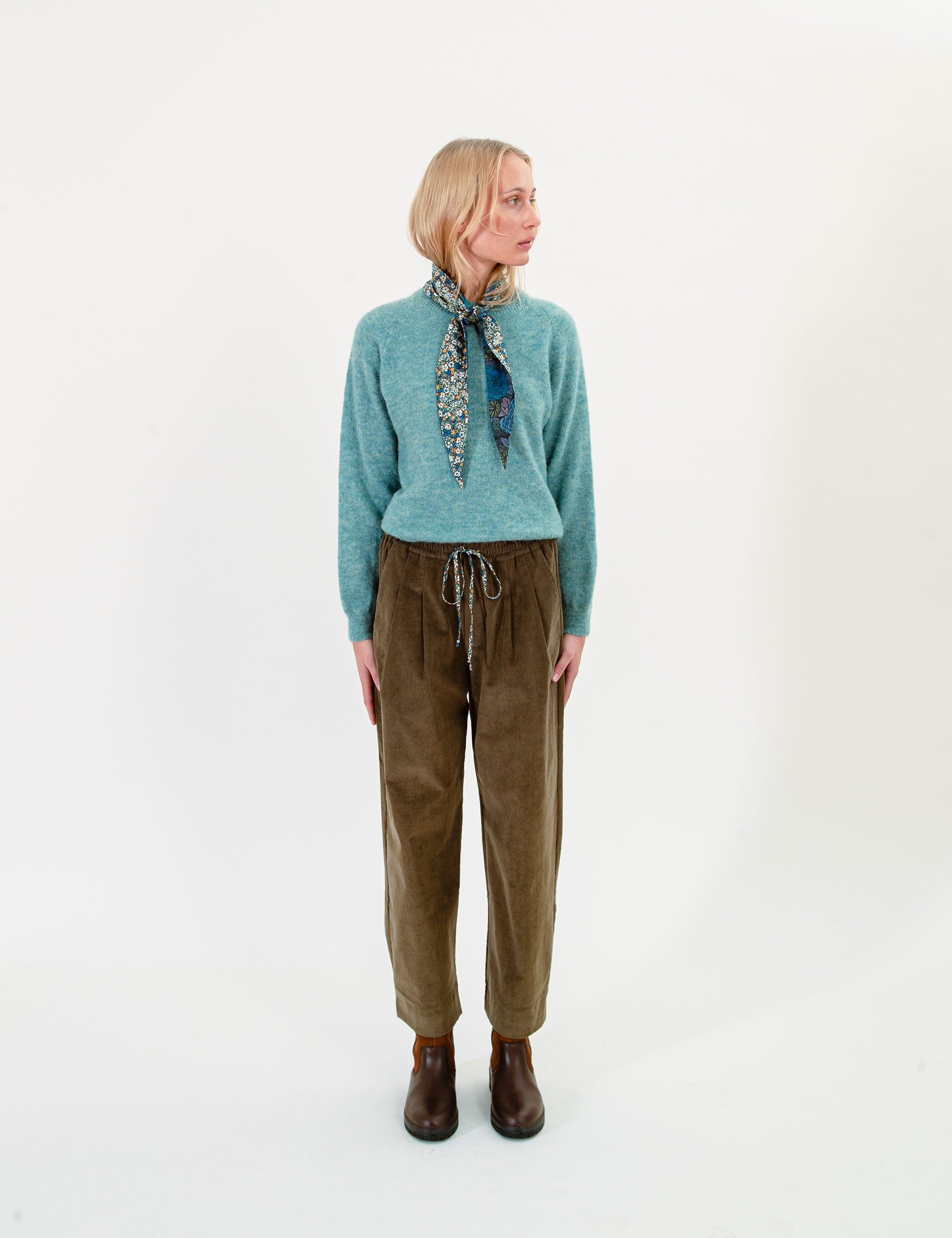 Woman wearing a teal sweater and olive green corduroy pants with a patterned scarf on a white background