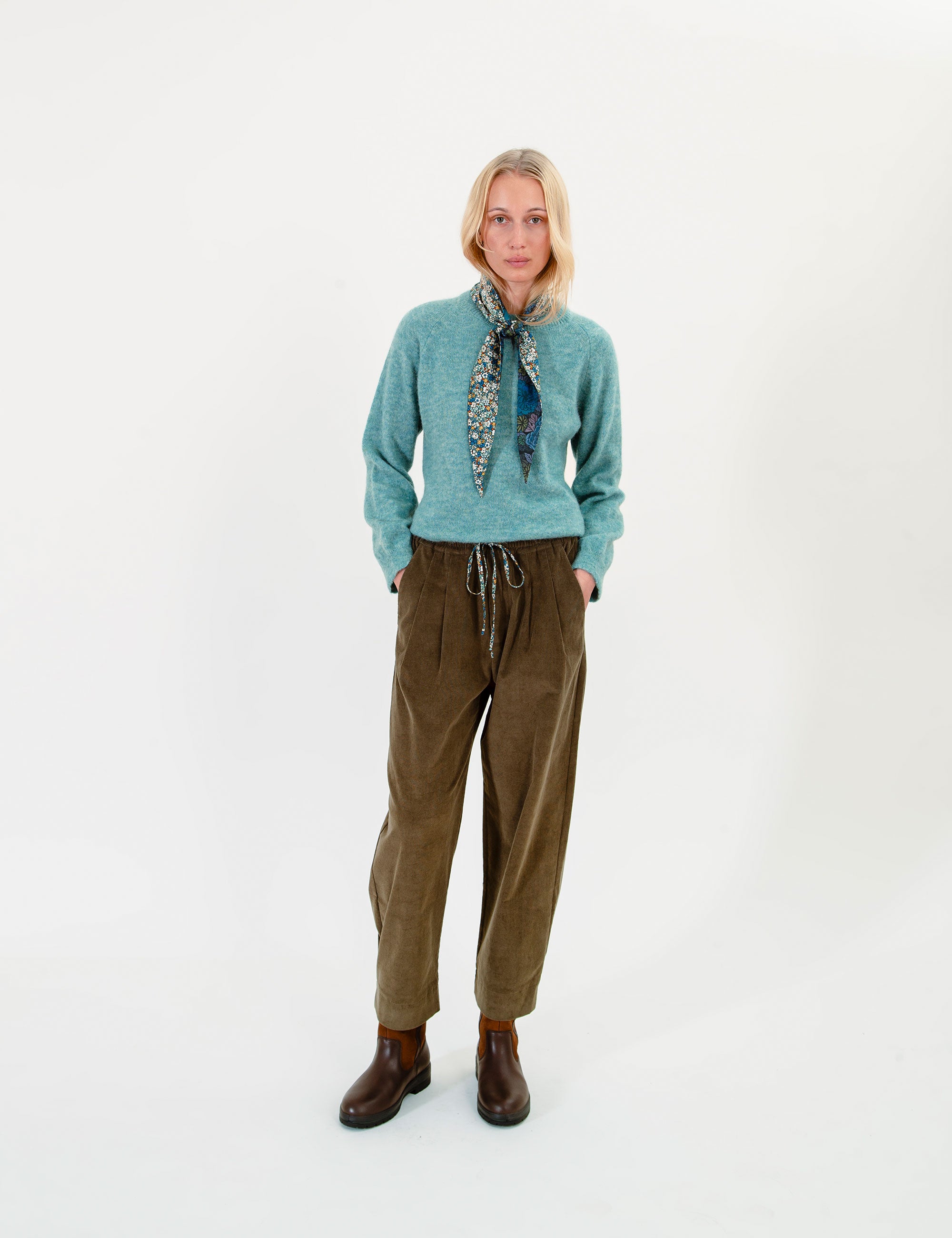 Woman wearing a teal sweater, olive green corduroy pants, and a patterned scarf on a white background